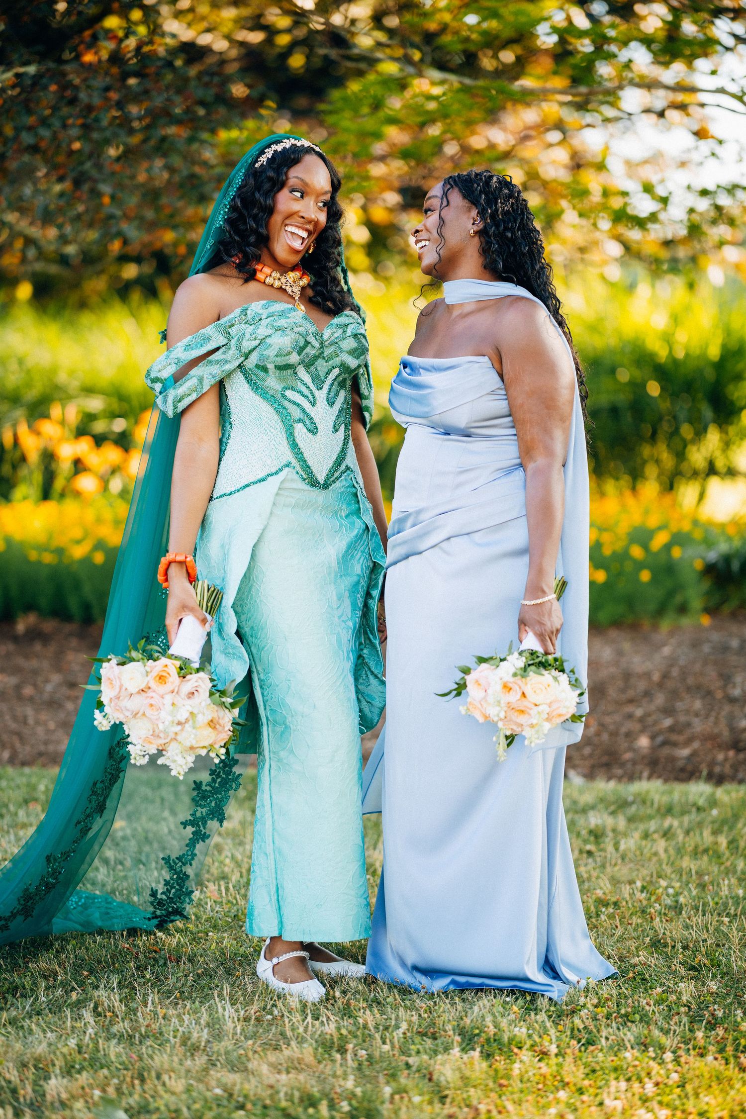 Two women in elegant gowns, one teal and one lavender, holding bouquets outdoors at a wedding.