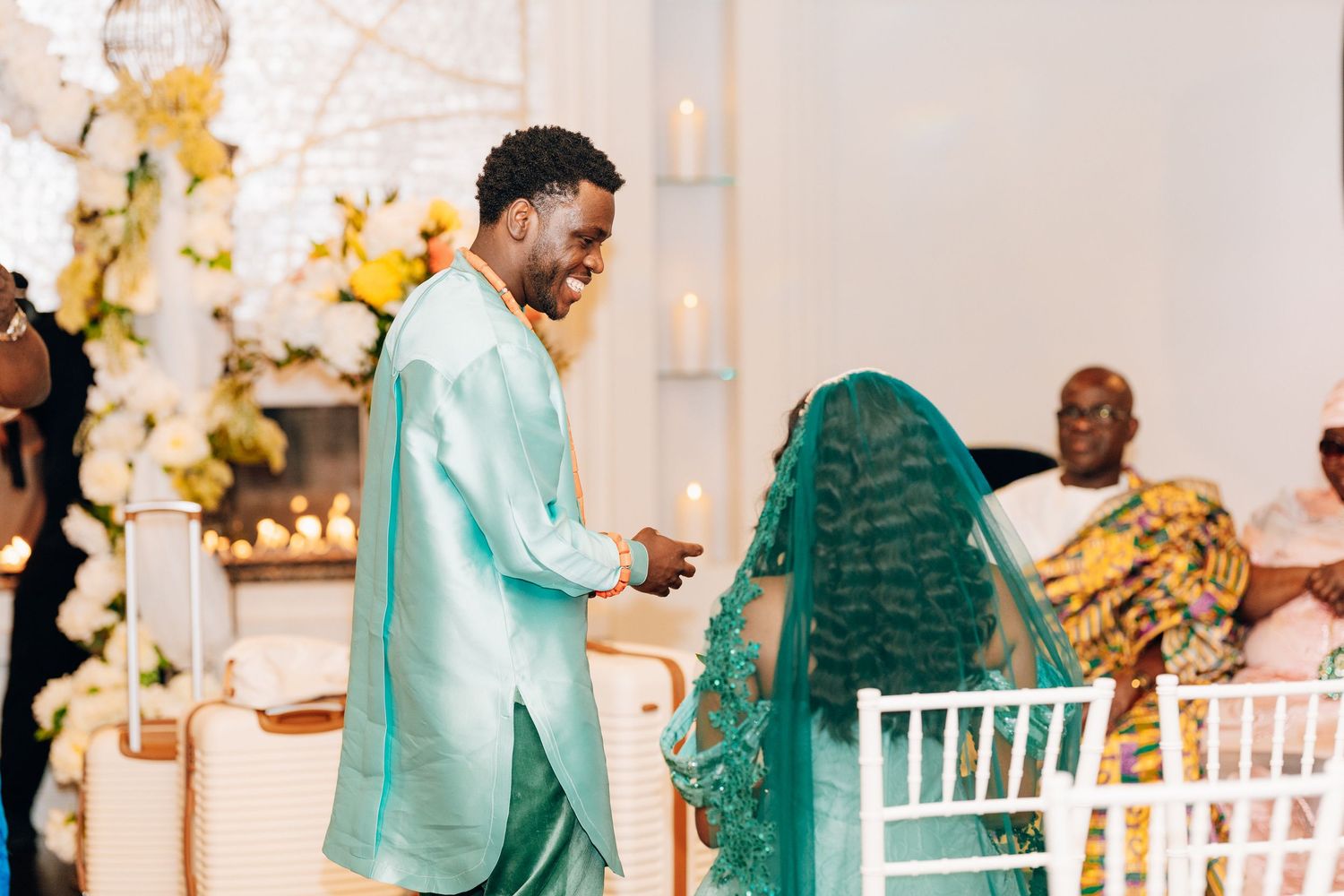 Groom in teal suit tenderly holding brides hand during traditional African wedding ceremony.