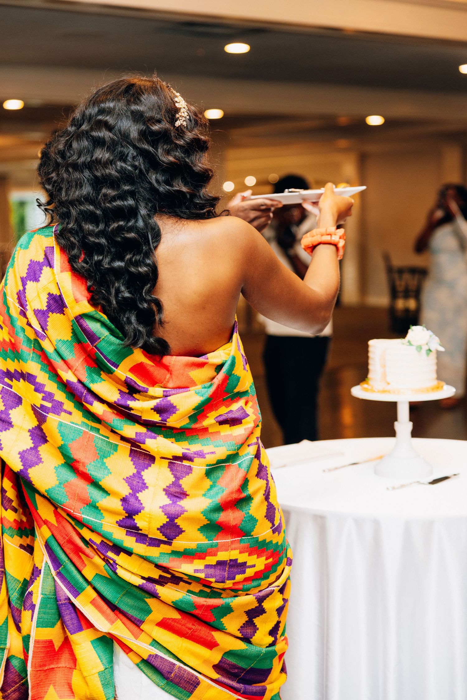 Bride in vibrant kente wrap dress preparing to cut wedding cake at reception.