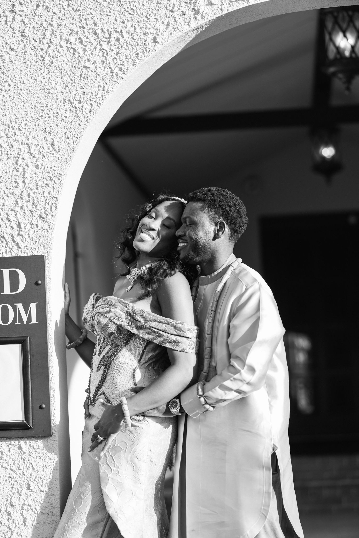 Black and white photo of a joyful couple embracing under an archway, dressed in elegant wedding attire.