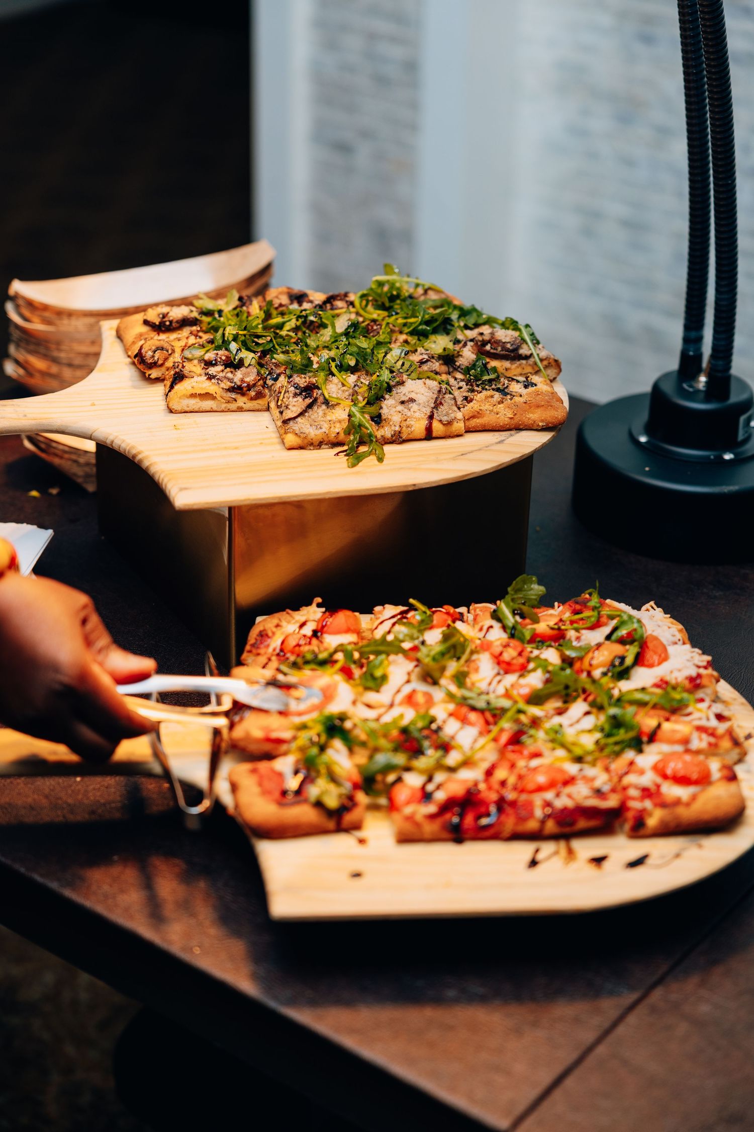 A hand serving slices of flatbread pizza topped with fresh herbs from wooden boards at a catering display.