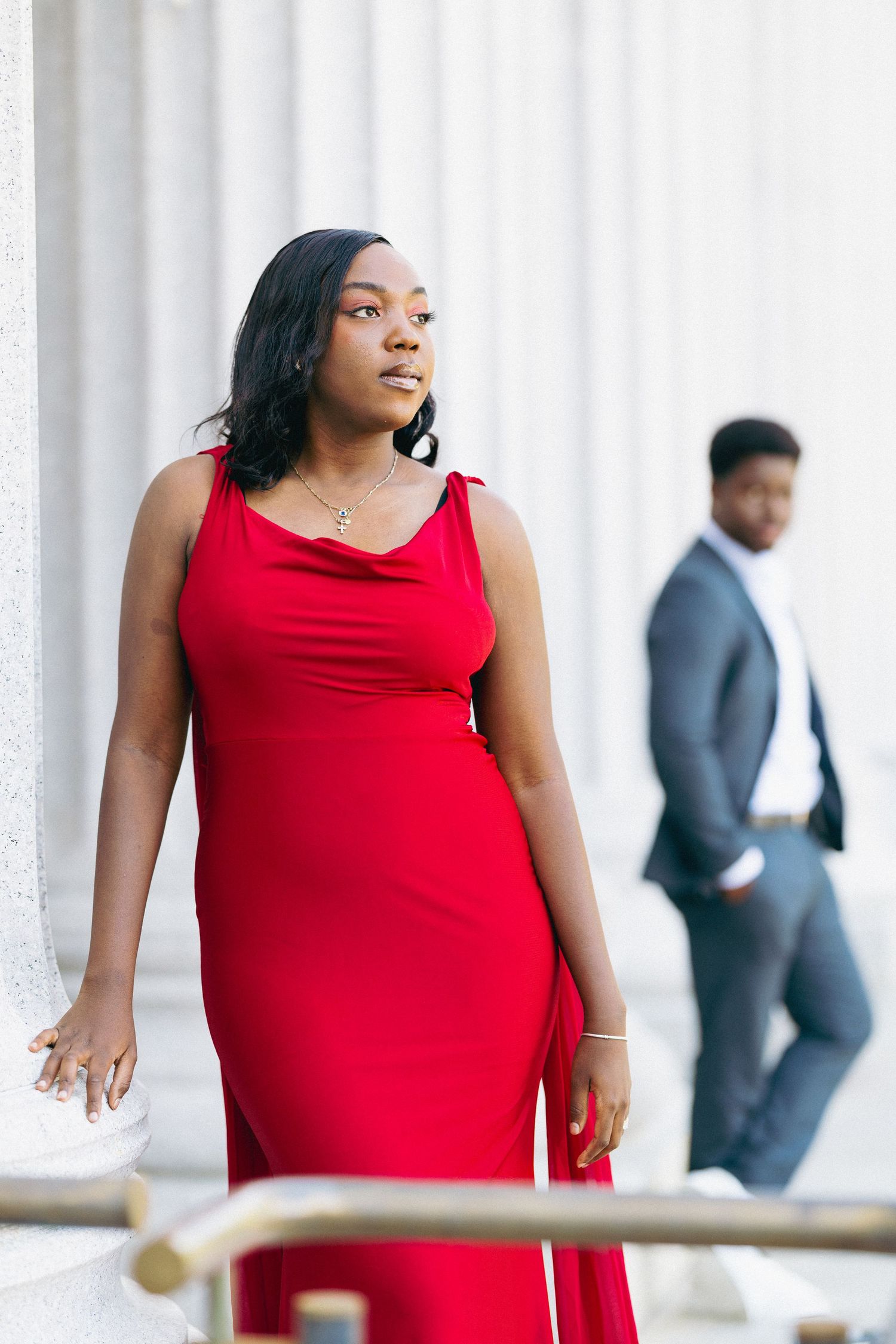 A woman in a red gown stands elegantly on steps near columns, with a man in a suit in the background.