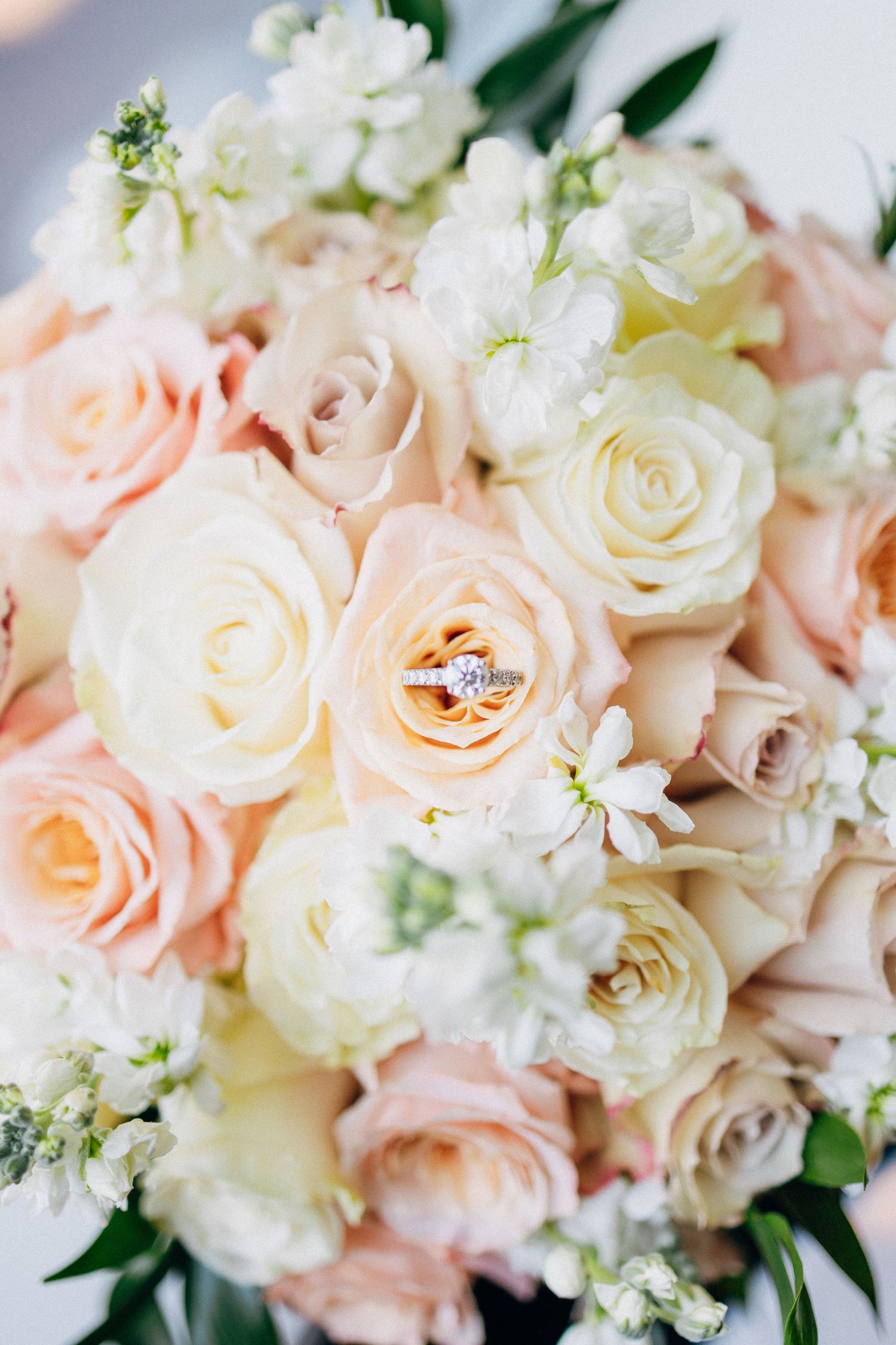 Wedding rings rest on a bridal bouquet of blush pink, peach, and white roses with delicate white blooms.