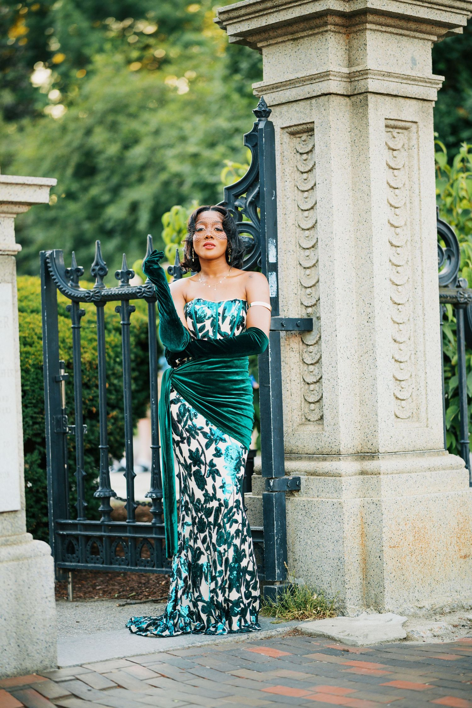 Elegant woman in green velvet floral gown with long gloves at iron gate.