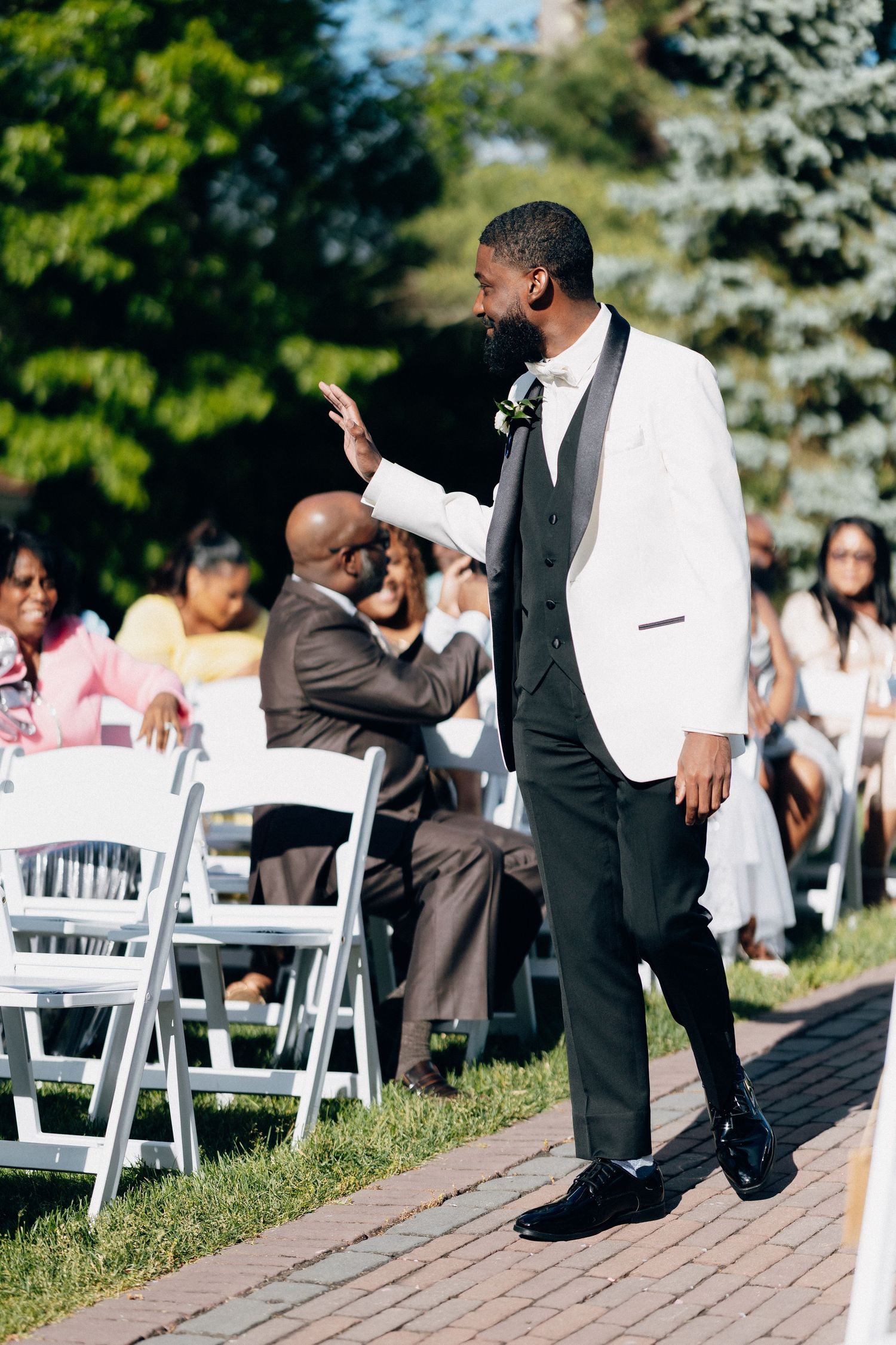 Groom in white tuxedo jacket waves to guests at outdoor wedding ceremony with white chairs.