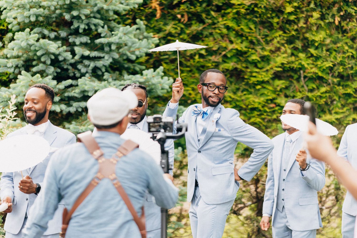 Groom in light blue suit celebrates joyfully with groomsmen in garden before wedding ceremony.