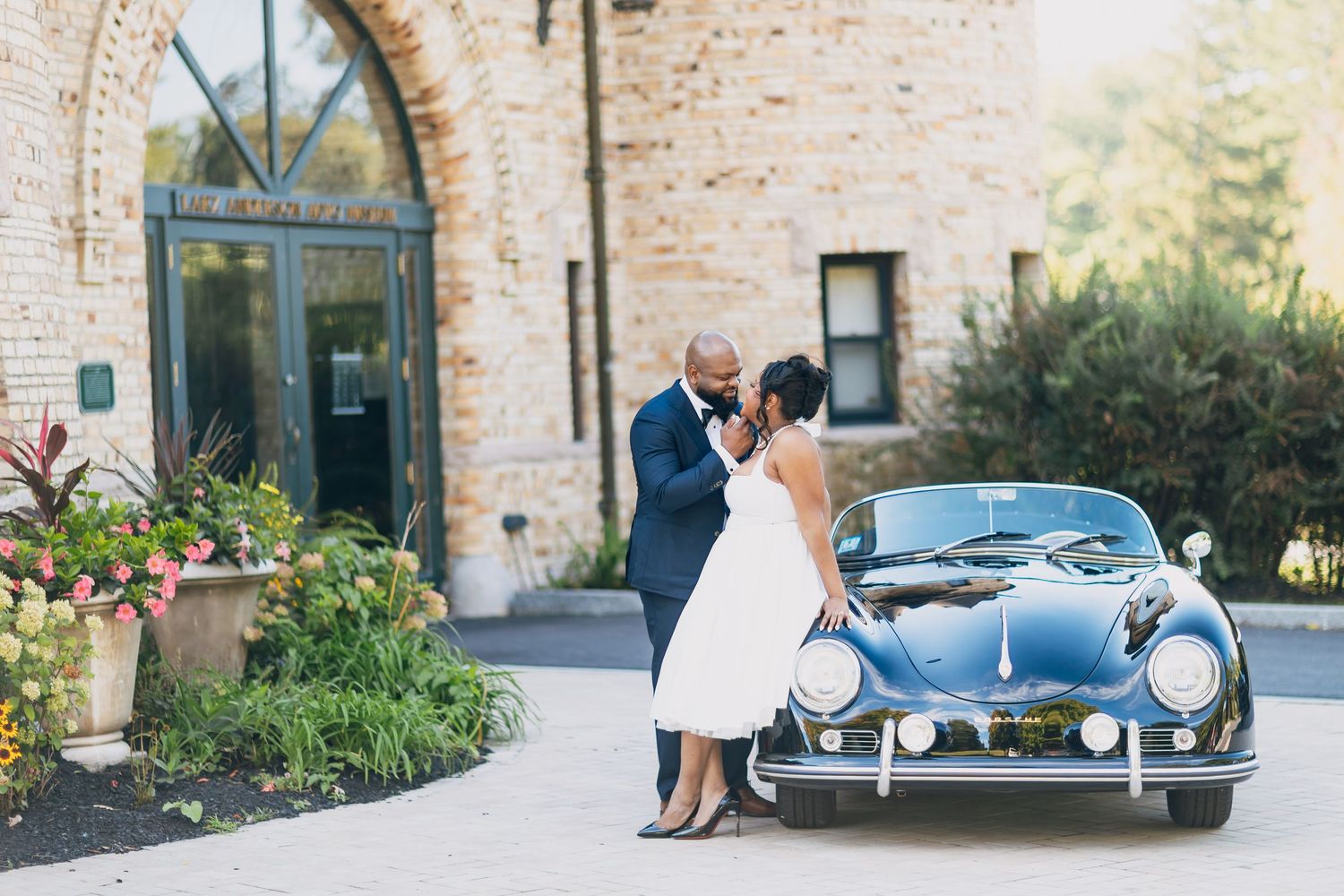 A couple shares a romantic kiss beside a vintage black Porsche outside a stone building on their wedding day.