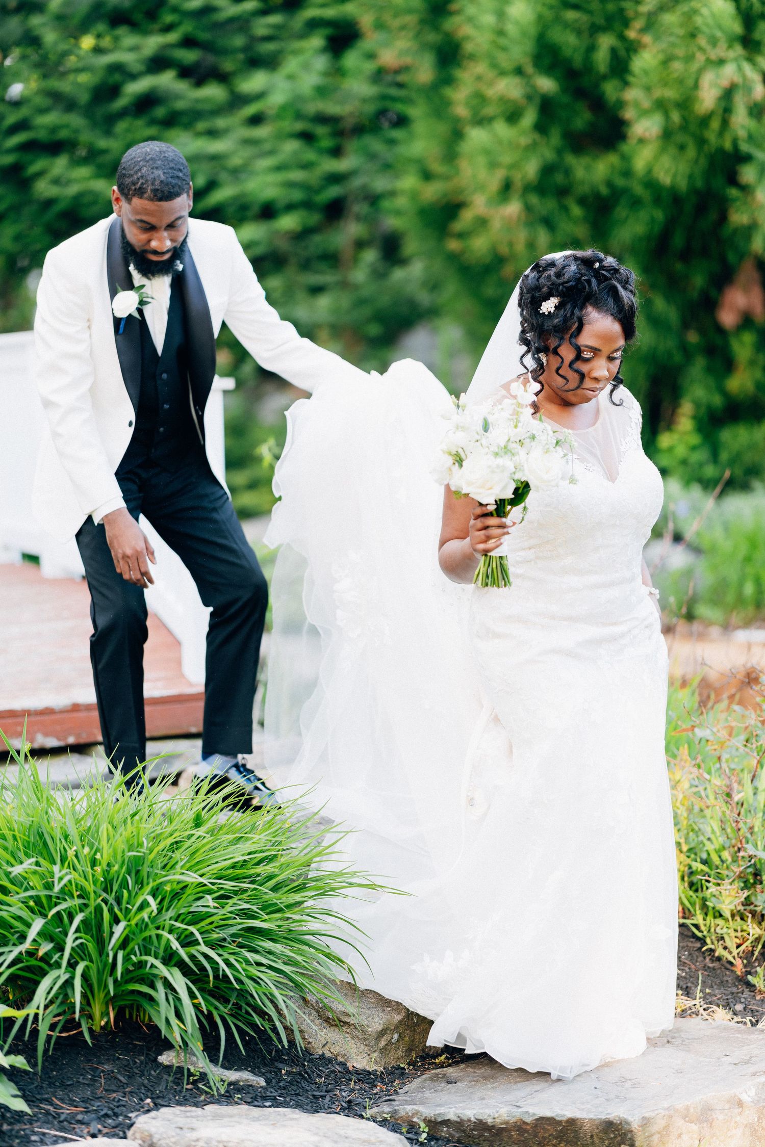 Groom in white tuxedo helps bride in white gown navigate stone steps in lush garden setting.
