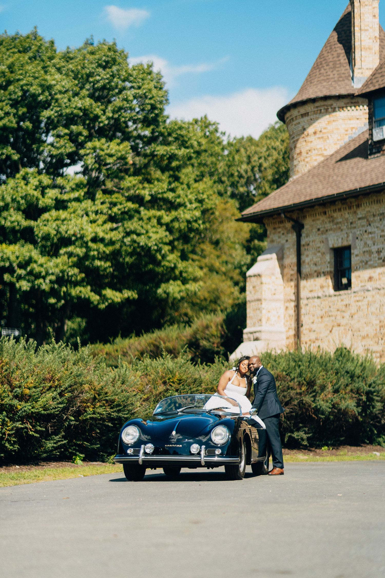 Wedding couple sharing a romantic kiss beside a vintage black Porsche 356 near a stone building.
