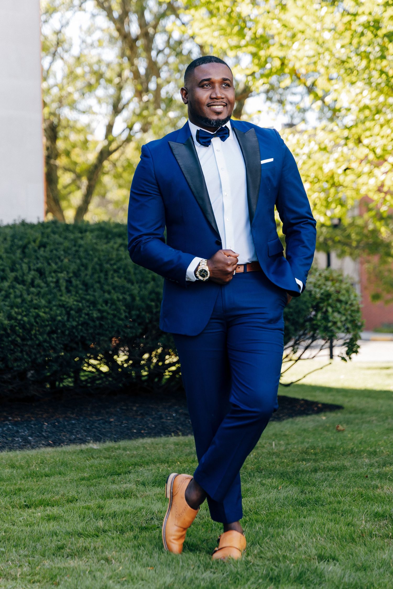 Stylish groom in navy blue tuxedo with bow tie and tan shoes poses confidently outdoors on green lawn.