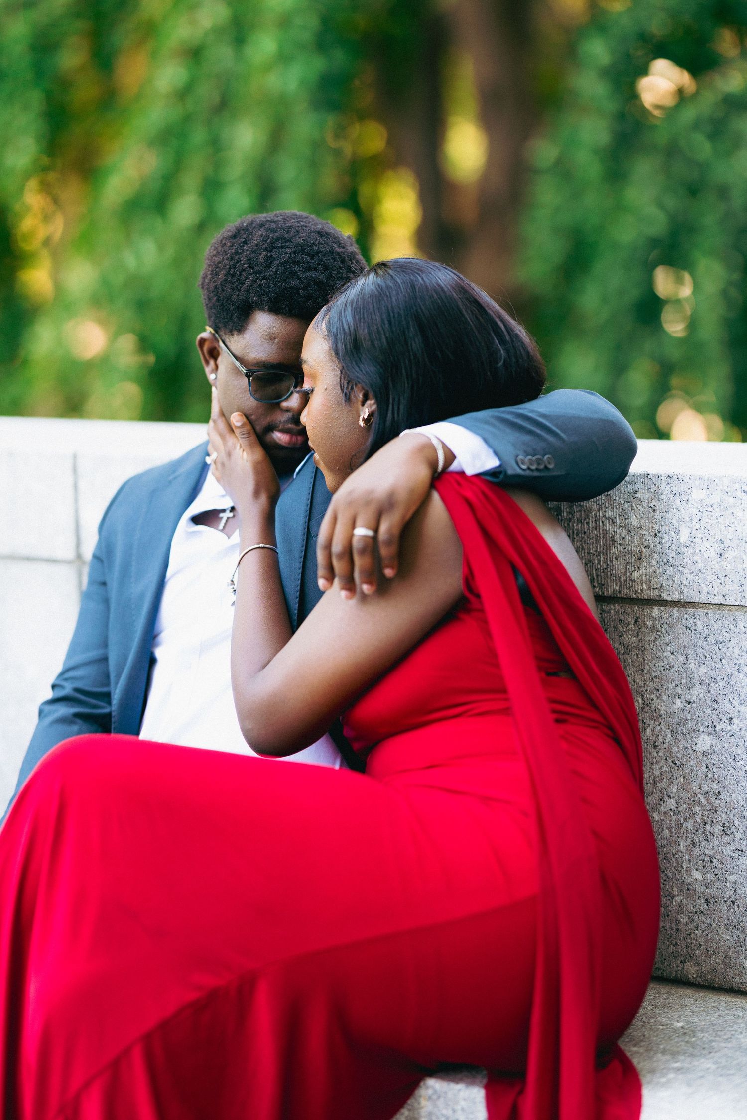 A couple in formal wear shares a romantic moment outdoors, woman in red gown rests on mans shoulder.