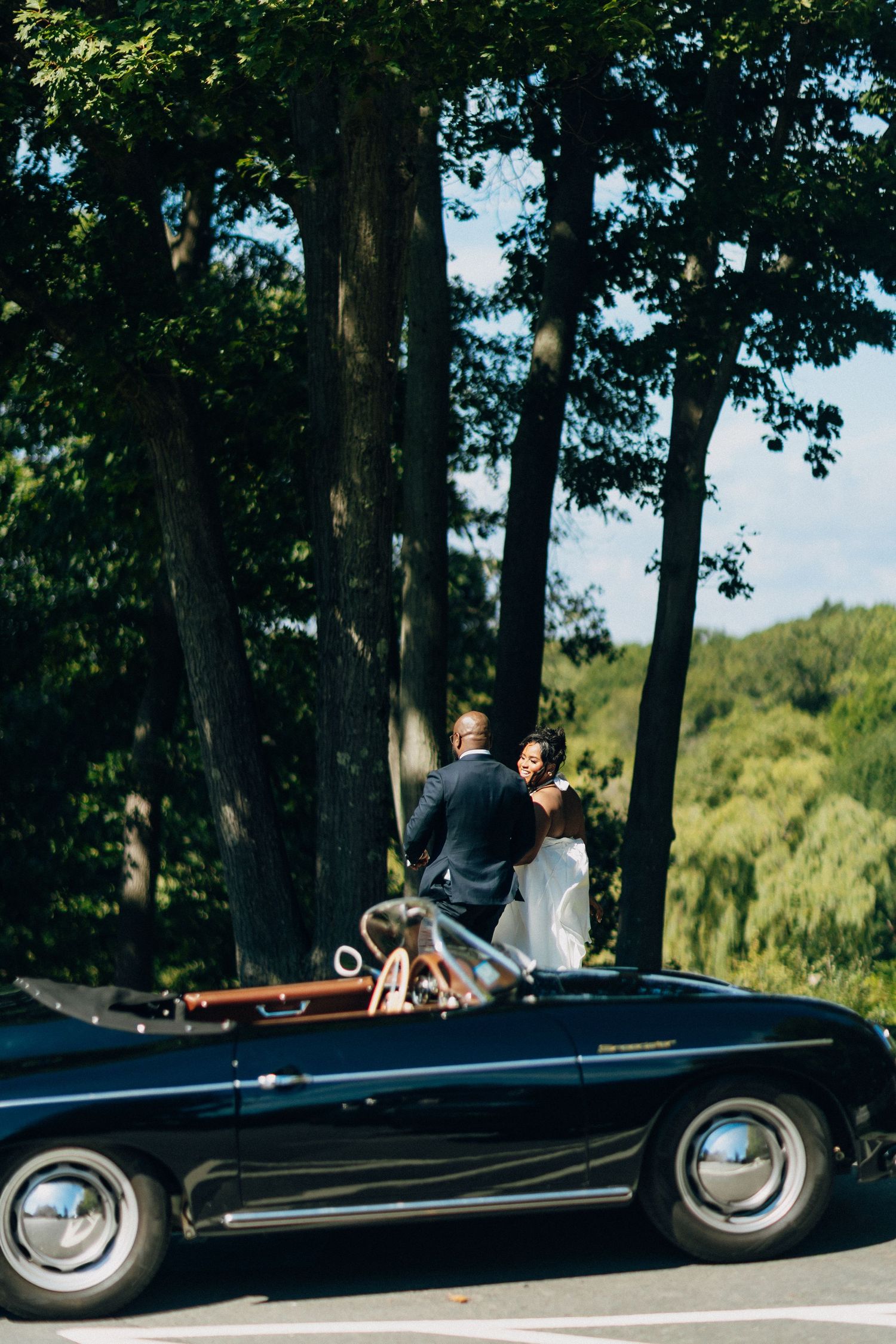 Bride and groom stand together in a classic black convertible surrounded by tall trees.