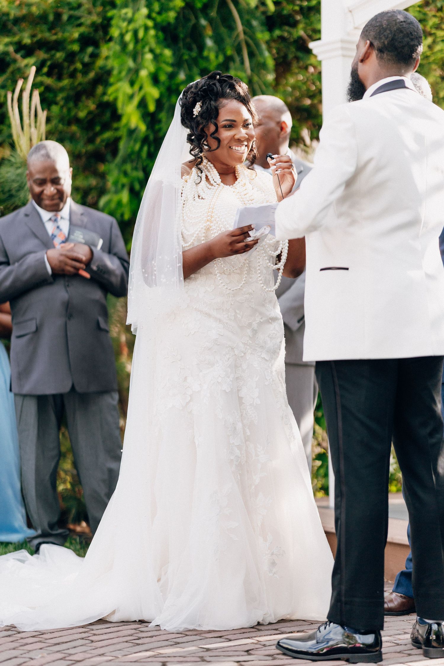 Smiling bride in lace gown reads vows to groom during outdoor wedding ceremony.