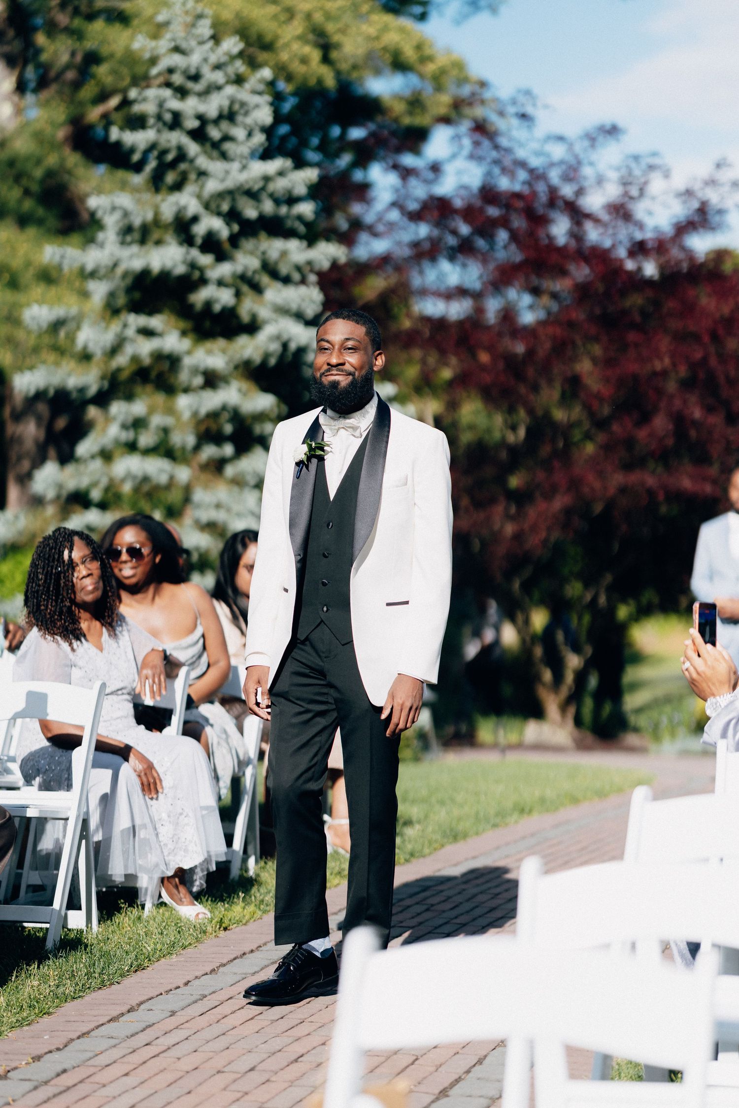 Smiling groom in white tuxedo jacket walks down outdoor wedding aisle with seated guests and green trees in background.