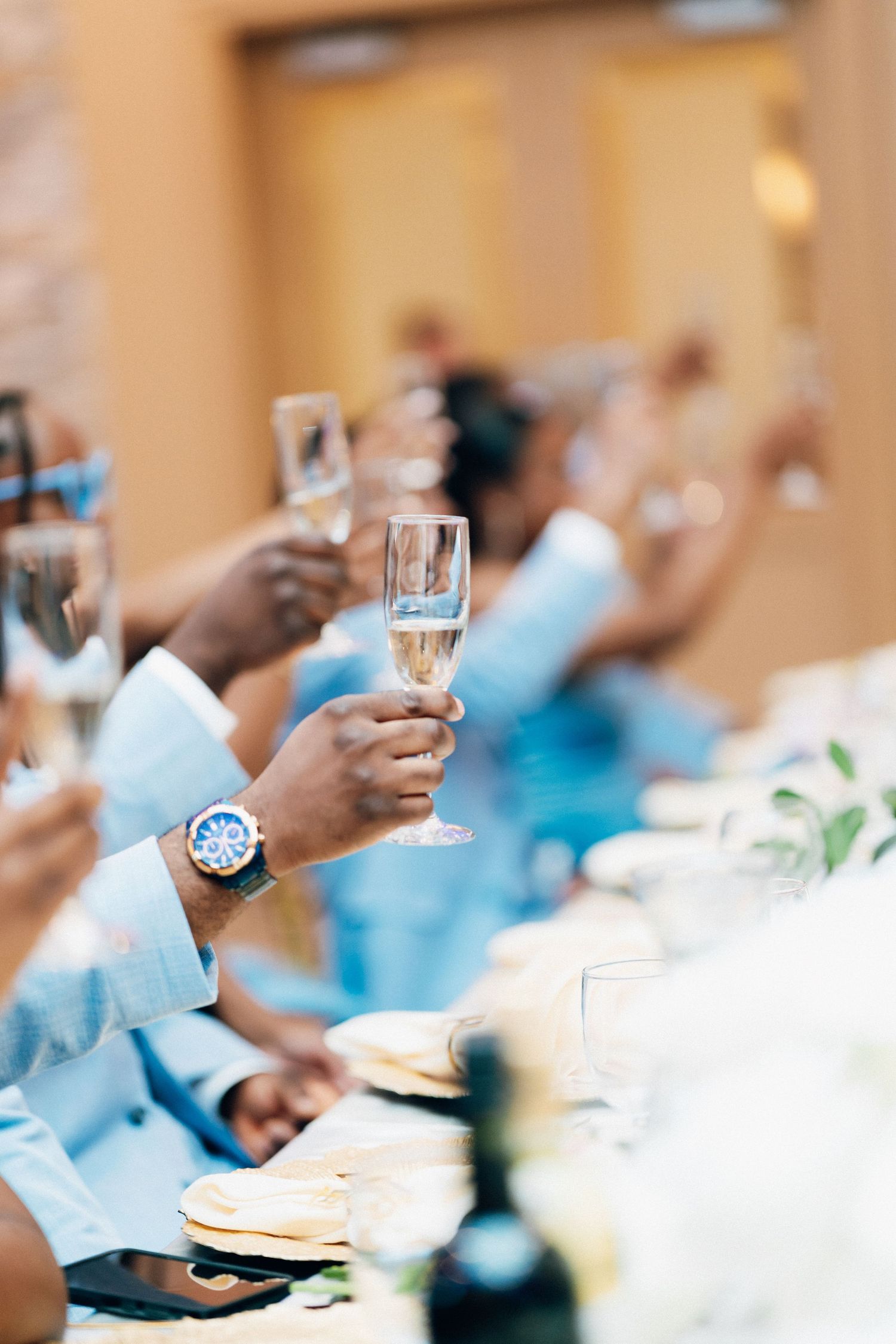 Groomsmen in light blue suits raising champagne glasses in a toast at a wedding reception dinner table.
