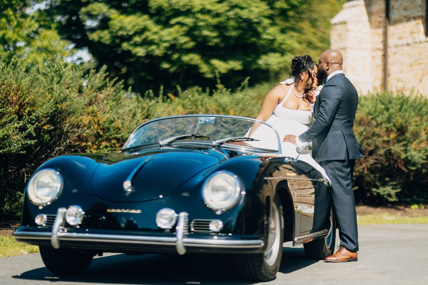 Bride and groom standing close together in front of a classic black Porsche convertible outdoors.