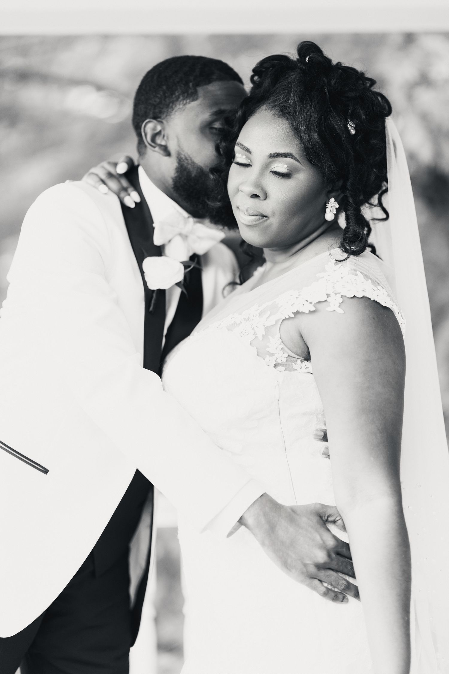 Black and white wedding photo of groom kissing bride on cheek while embracing her from behind in elegant attire.