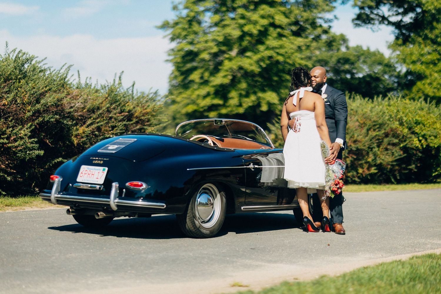 Newlywed couple embracing behind a classic black vintage convertible on a tree-lined driveway.