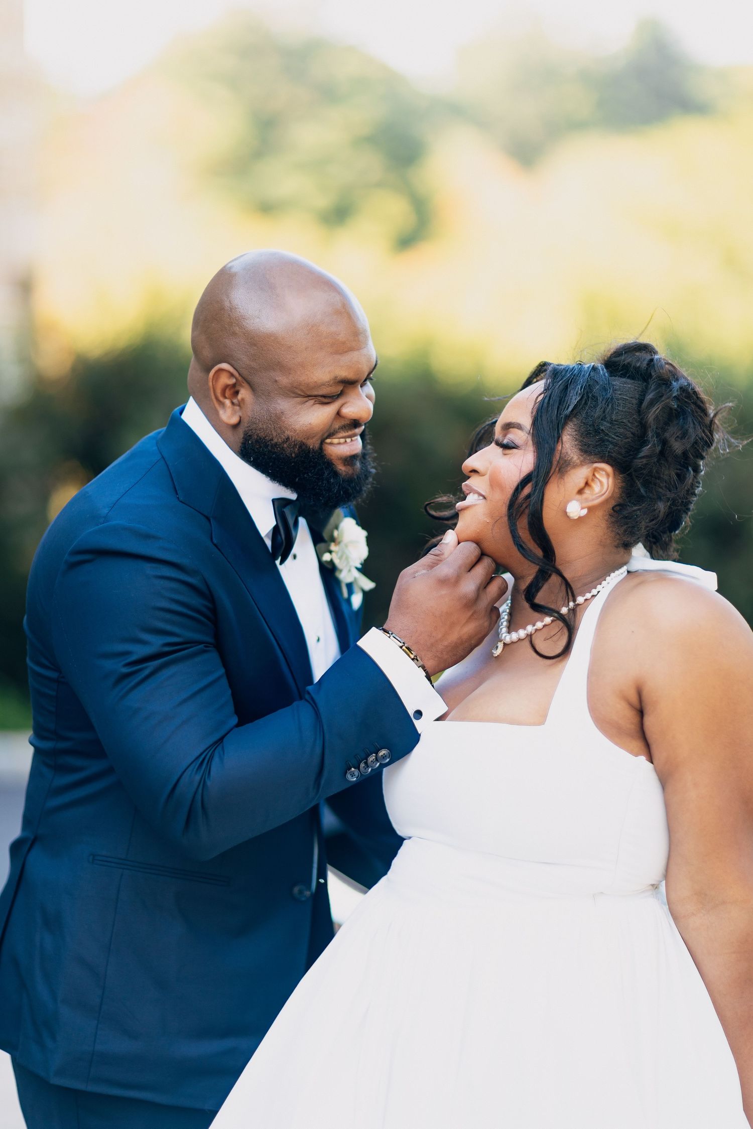 Joyful newlyweds sharing a tender moment outdoors, groom in navy suit gently touching brides chin as they laugh together.