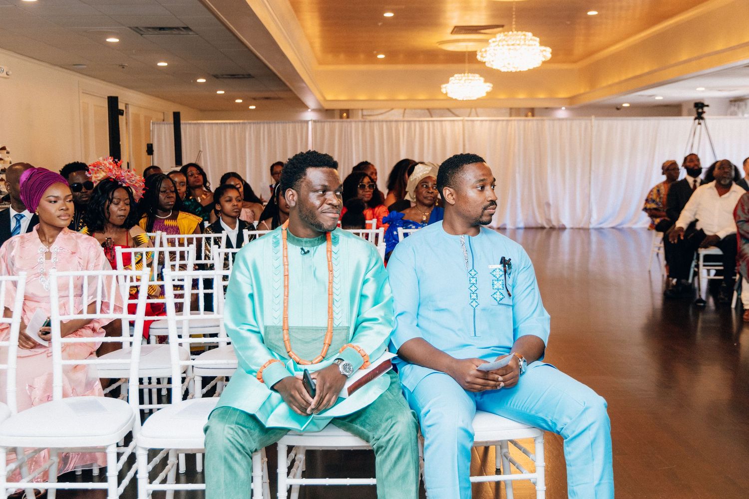 Two men in traditional African attire seated at an elegant wedding ceremony venue with white chairs and draped decor.