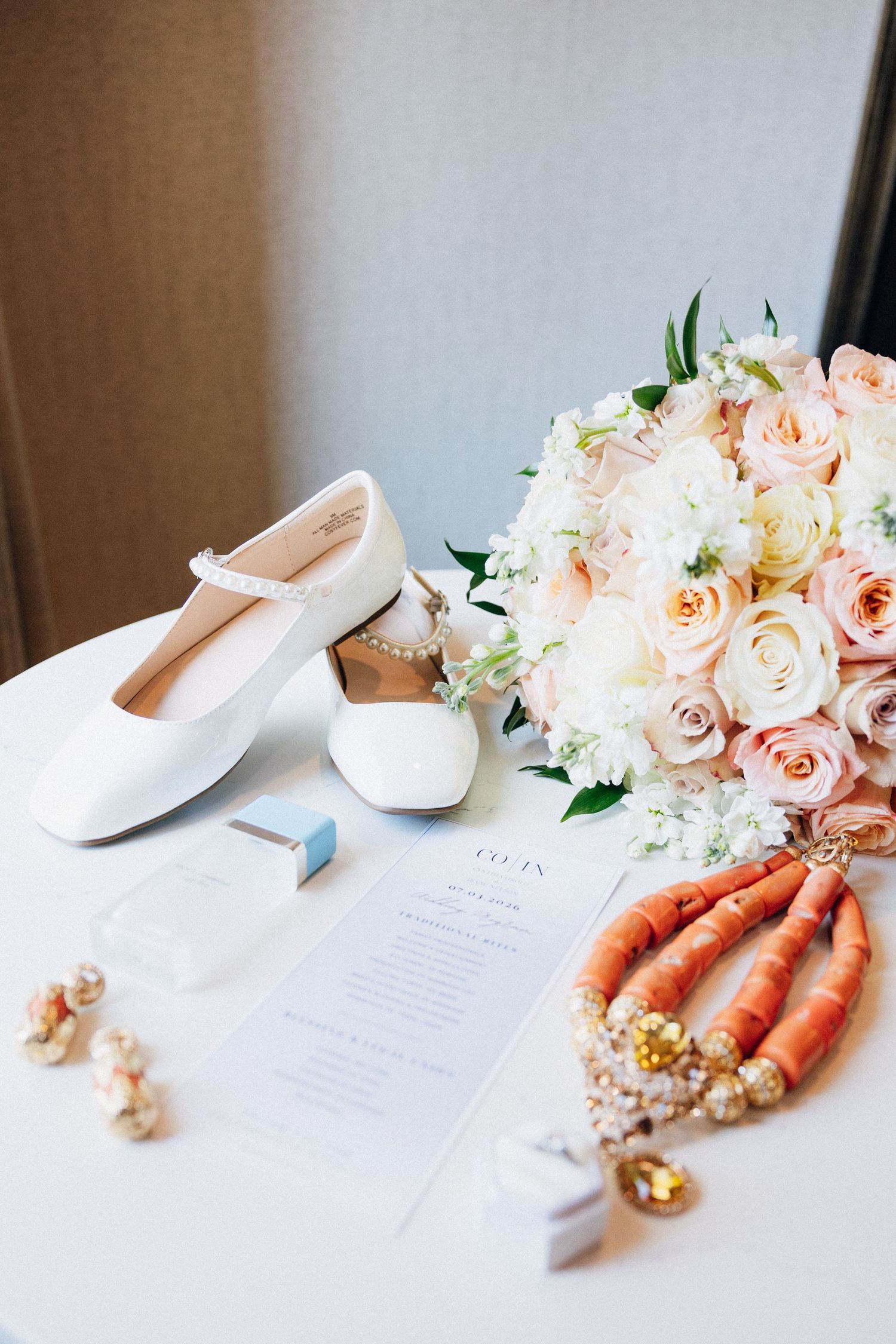 White bridal flats, coral beaded necklace, pearl earrings, pink rose bouquet, and wedding invitation flatlay on white table.