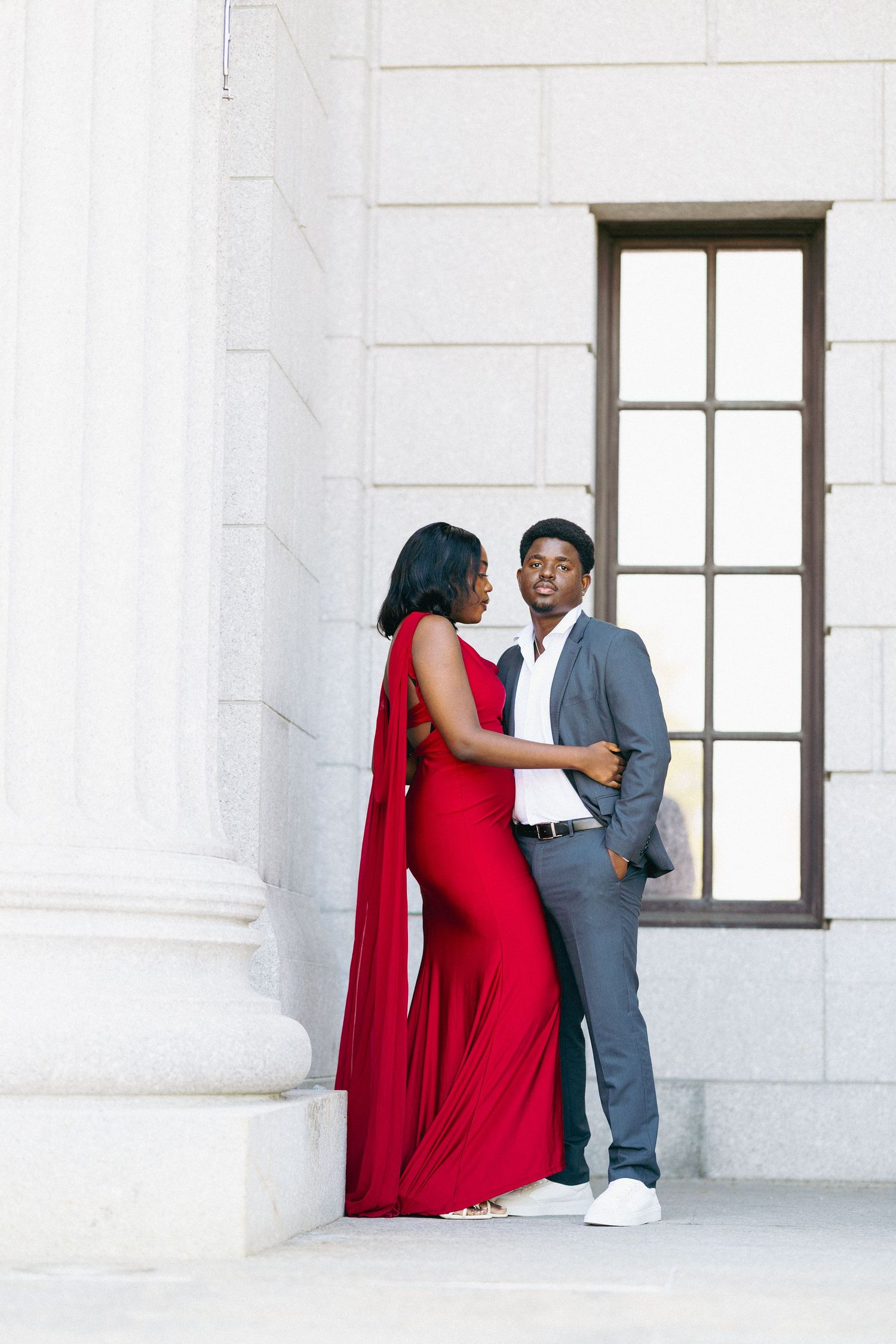Couple posing elegantly, woman in red cape gown, man in gray suit, against white stone columns and window backdrop.