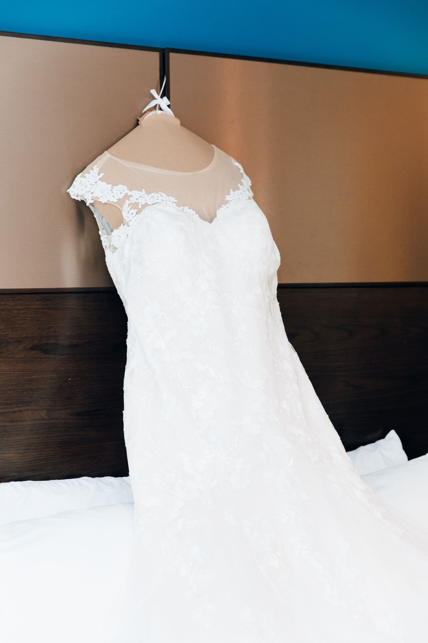 Elegant white lace wedding dress with illusion neckline hanging on a hanger in a hotel room.