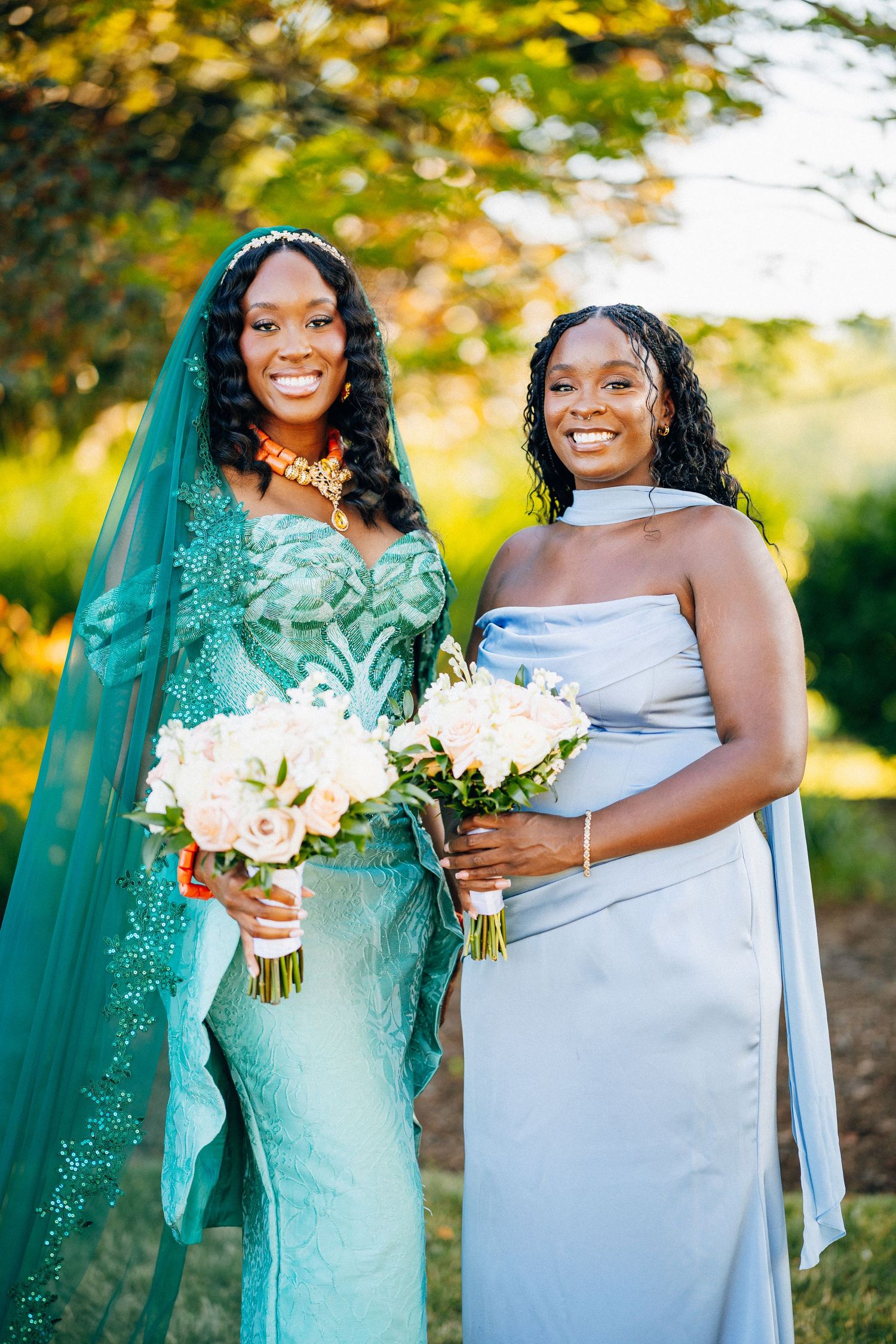 Two smiling women in elegant wedding attire holding bouquets outdoors, one in teal gown with veil, other in blue dress.