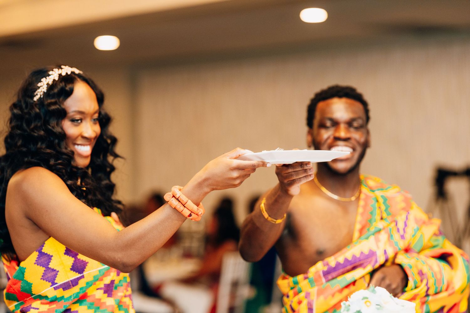 Couple in colorful Kente cloth attire sharing cake at a joyful African wedding celebration.