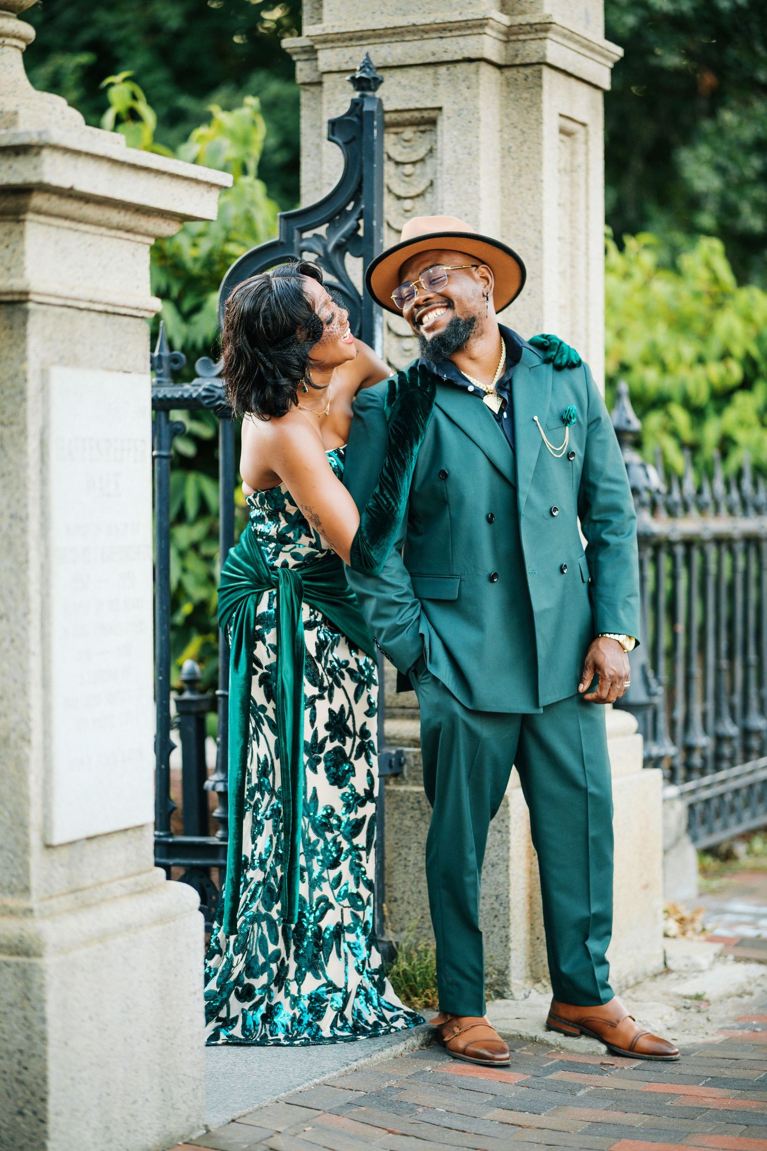 Joyful couple in matching green formal attire posing by ornate iron gate.