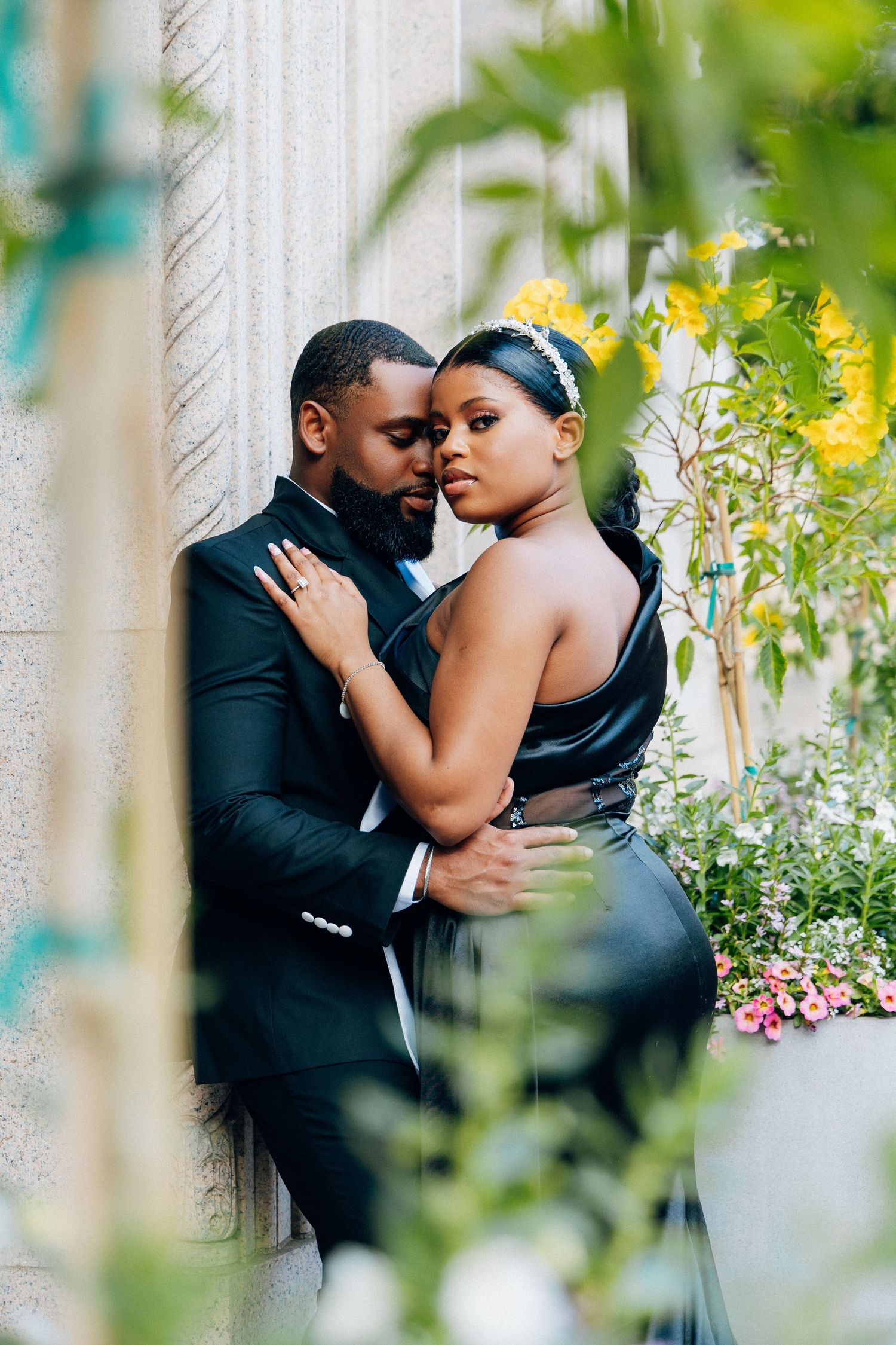 A couple in elegant black formal wear embraces romantically against a stone column surrounded by lush greenery and flowers.