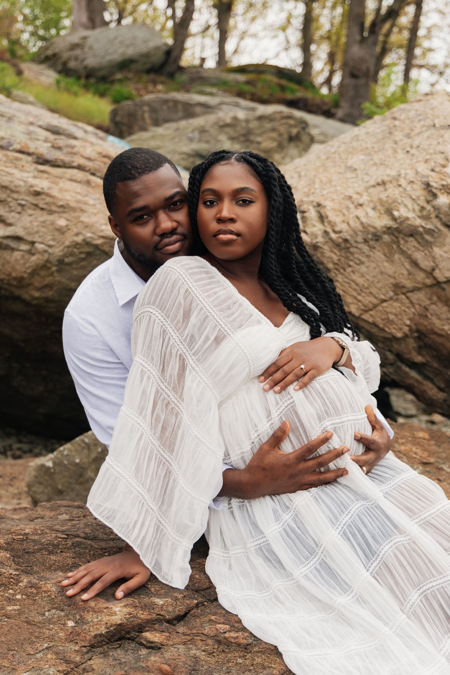 Pregnant Black couple in white outfits sitting on rocks outdoors, man embracing woman from behind during maternity photoshoot.