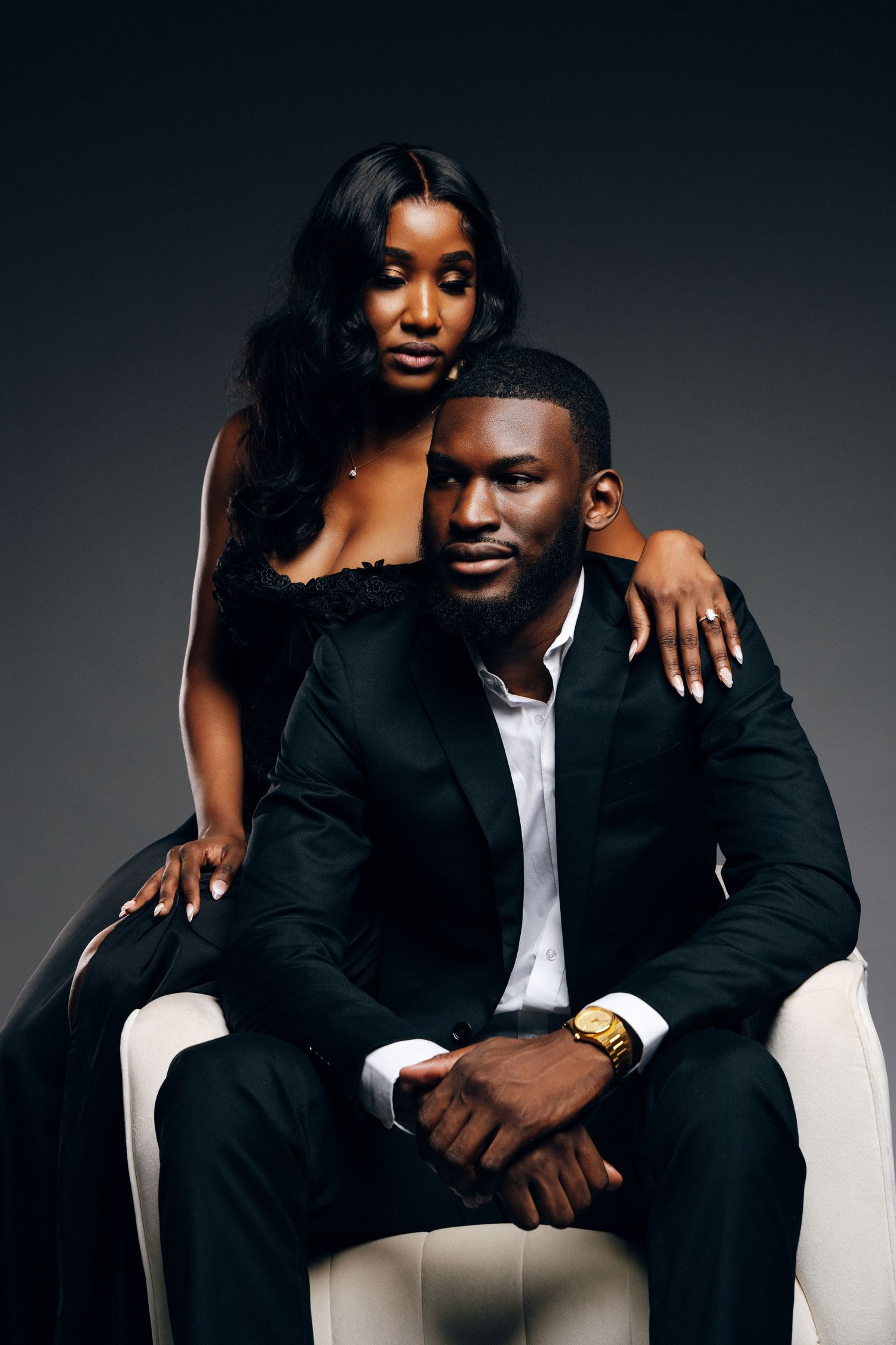 Elegant Black couple in formal attire, man seated in white chair wearing suit and gold watch, woman standing behind him.