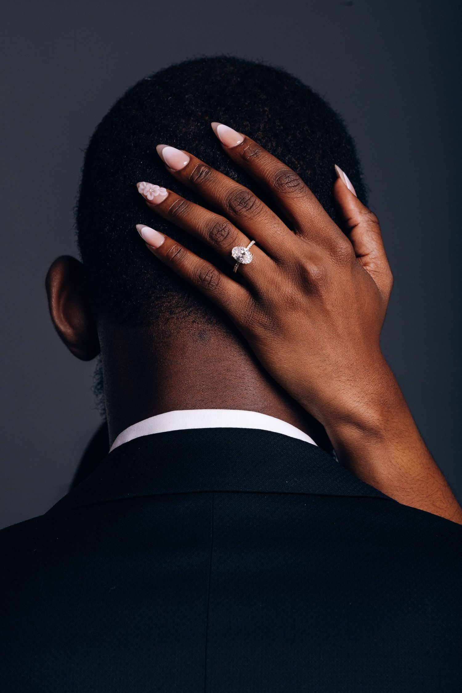 A woman's hand with a diamond engagement ring rests on the back of a man's head in a black suit.
