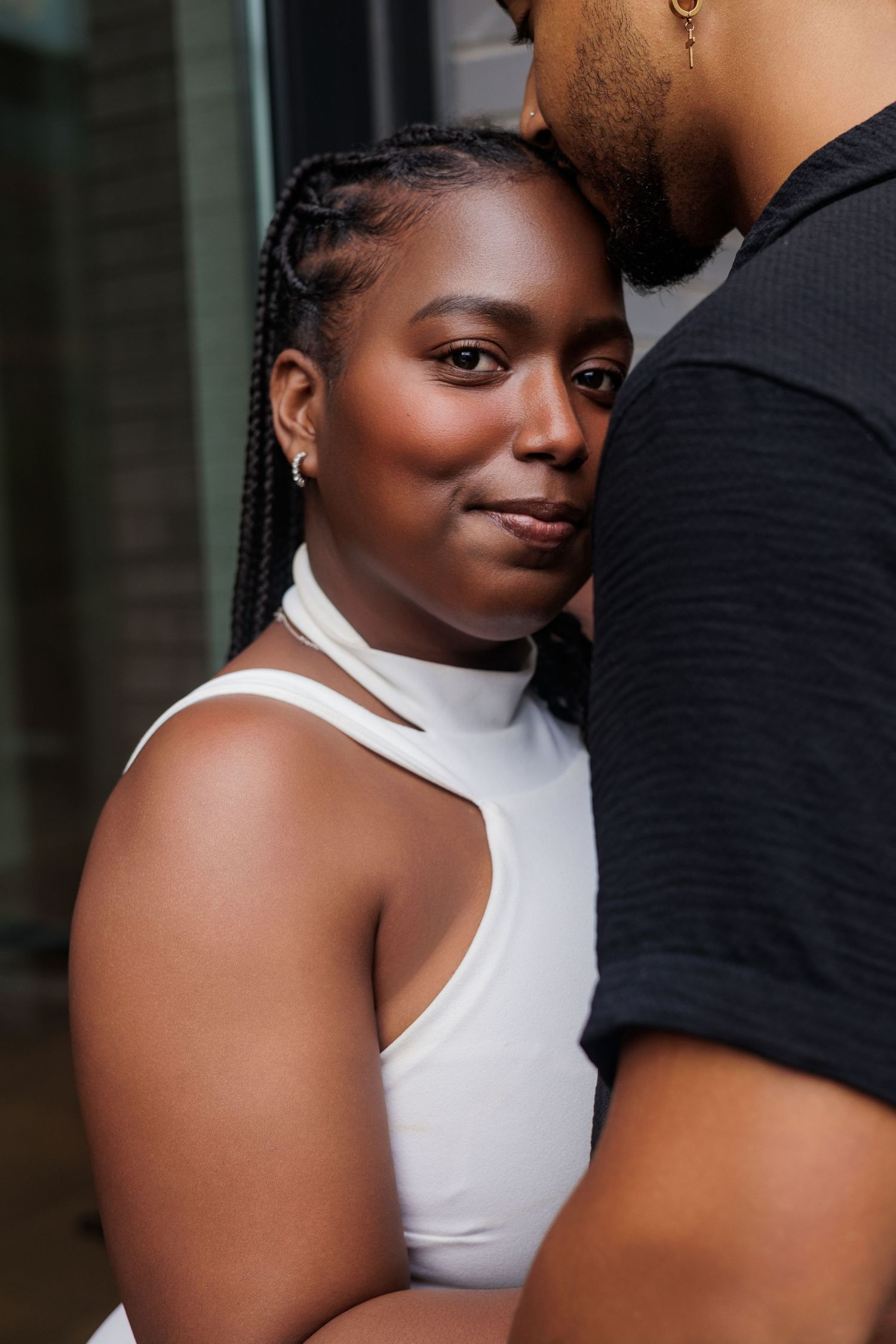 A woman in a white top leans against a man in a black shirt, smiling softly at the camera.