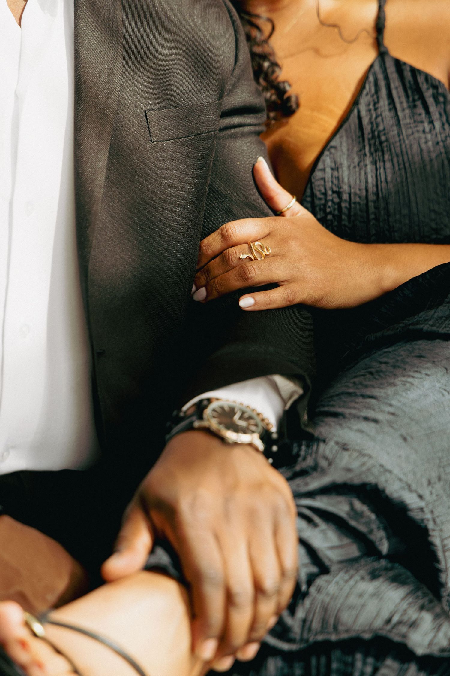 Close-up of a couple holding hands, woman wearing a gold ring and man wearing a luxury watch, both dressed in elegant dark attire.