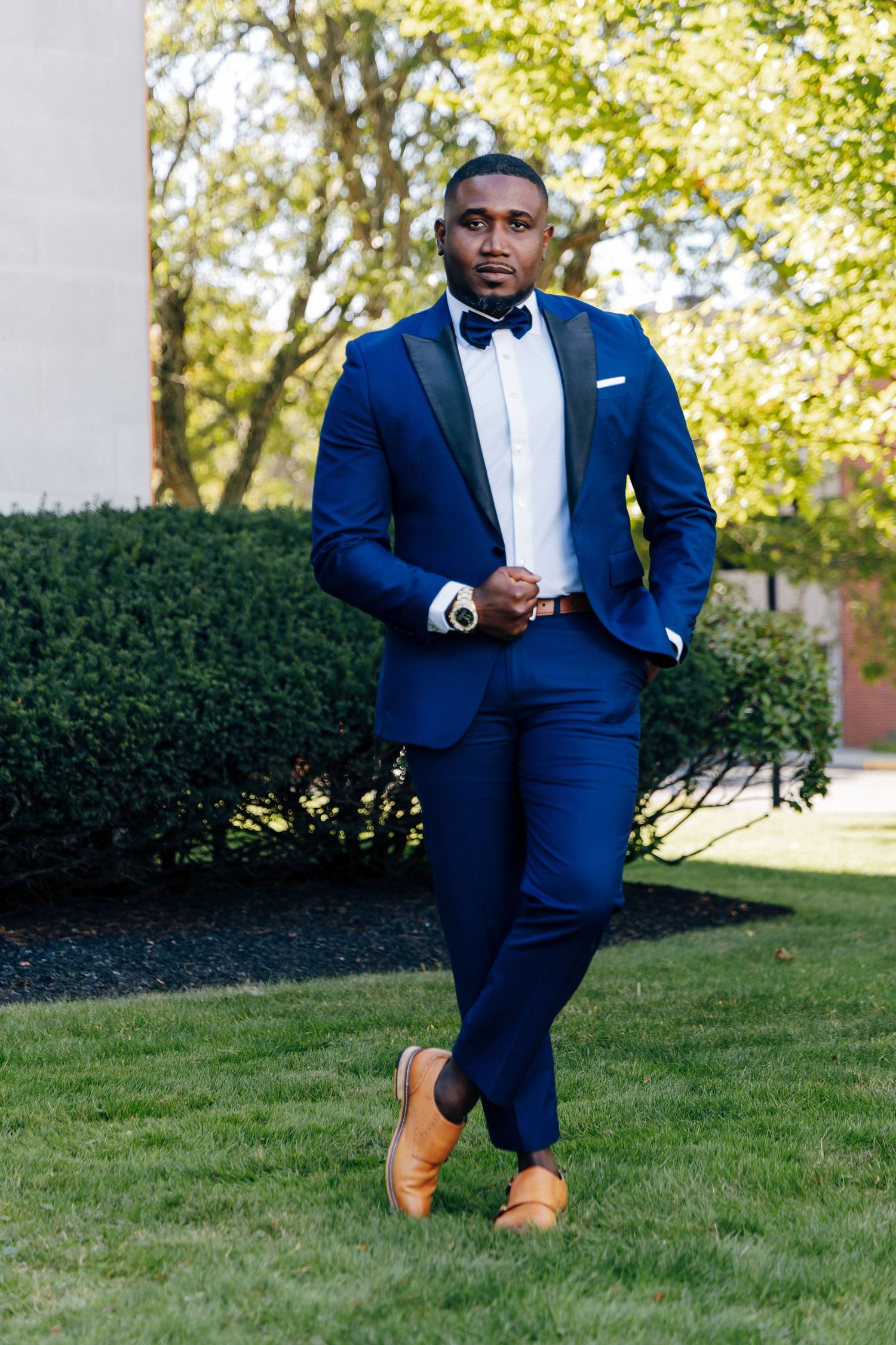Groom in navy blue tuxedo with bow tie and tan shoes posing outdoors on green lawn.