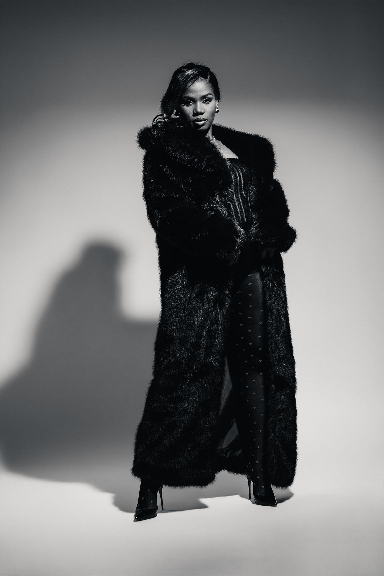 Black and white photo of a woman in a long black fur coat and heels posing confidently in a studio.