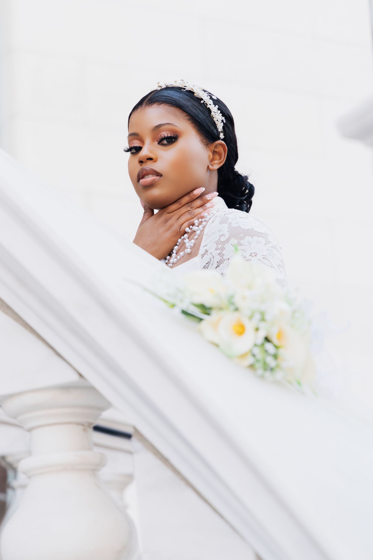 Elegant Black bride in lace wedding dress holding bouquet, wearing crystal headband, posing gracefully on white staircase.