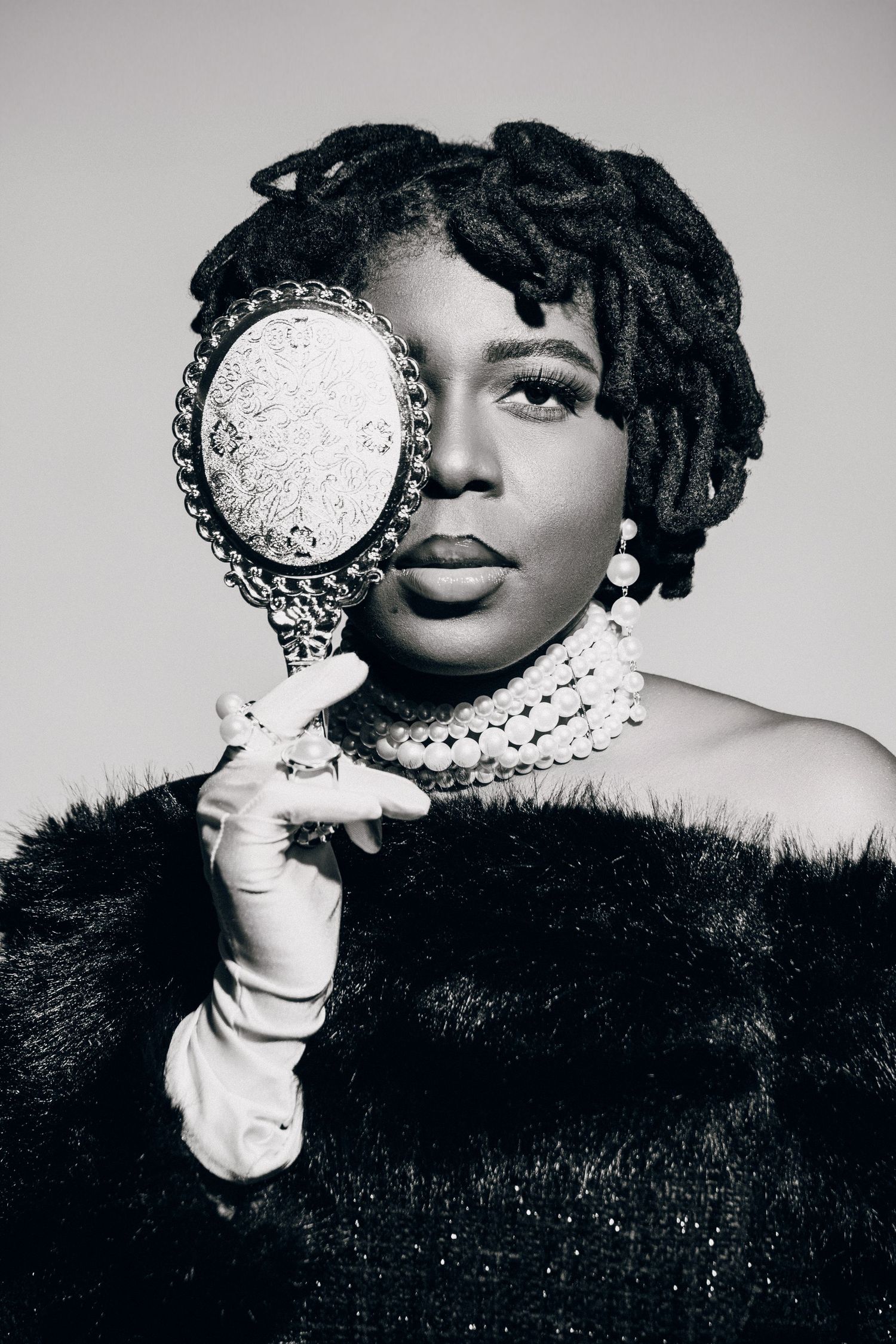 Black and white portrait of a woman with dreadlocks holding an ornate hand mirror, wearing fur coat and pearl necklace.