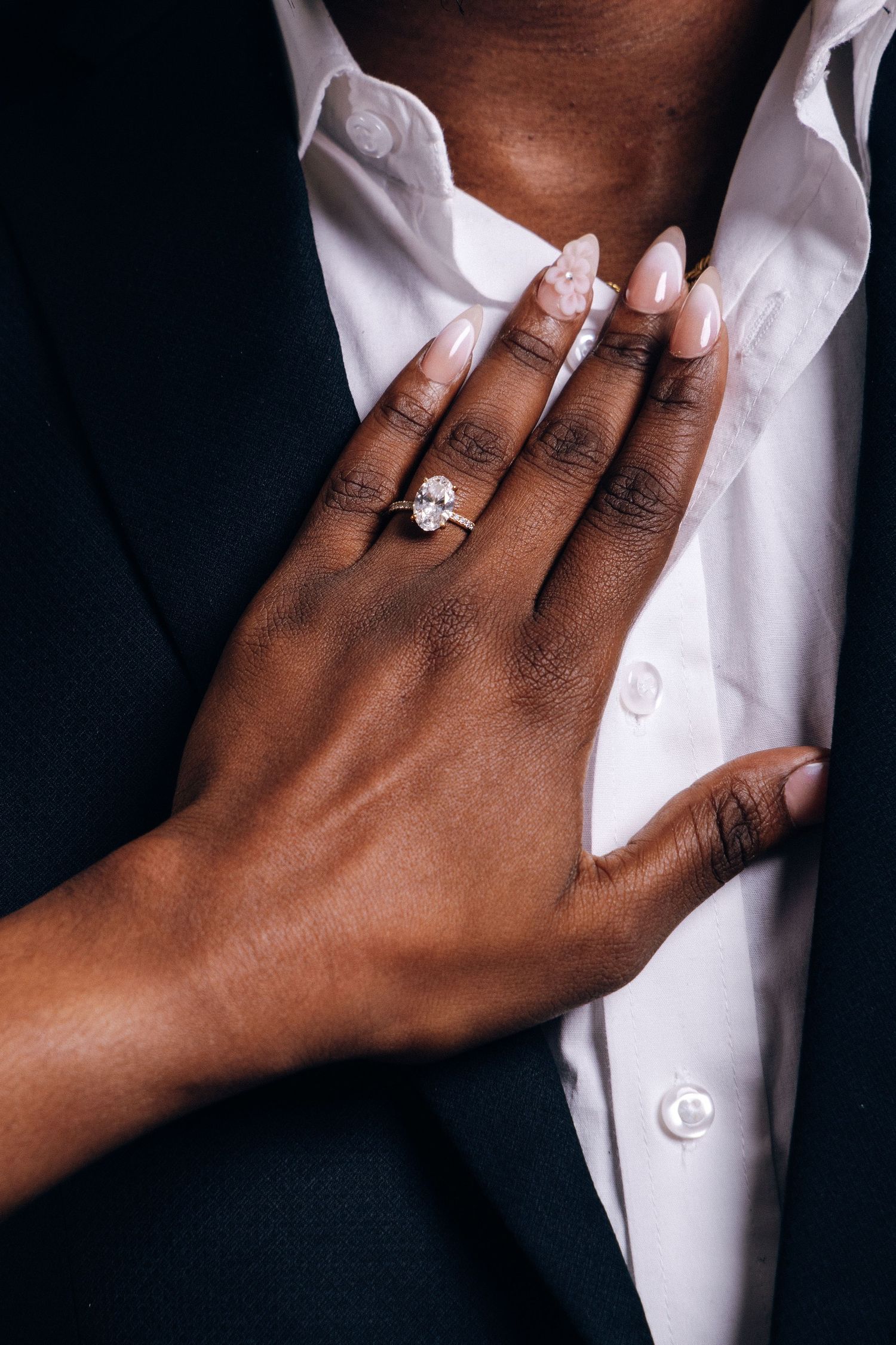 A woman's hand with nude nails displays a large diamond engagement ring while resting on a man's white dress shirt.