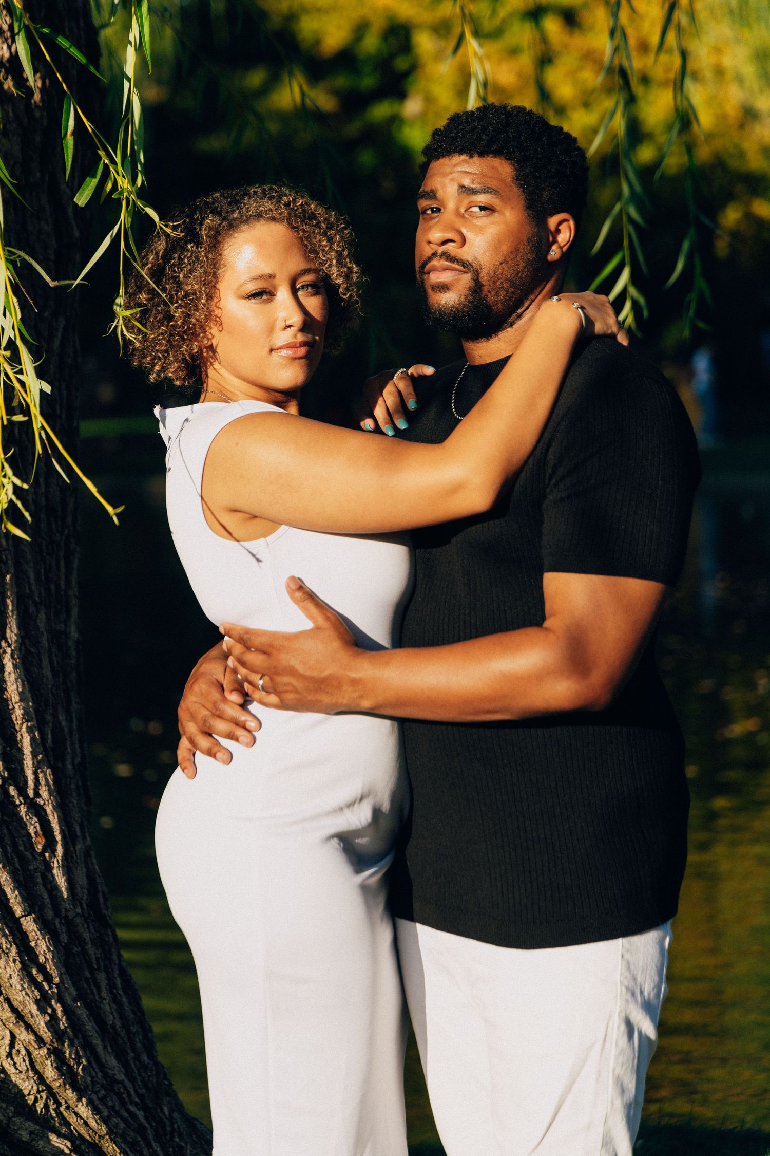 A couple embraces tenderly near a willow tree, the woman in white and man in black sharing an intimate moment outdoors.