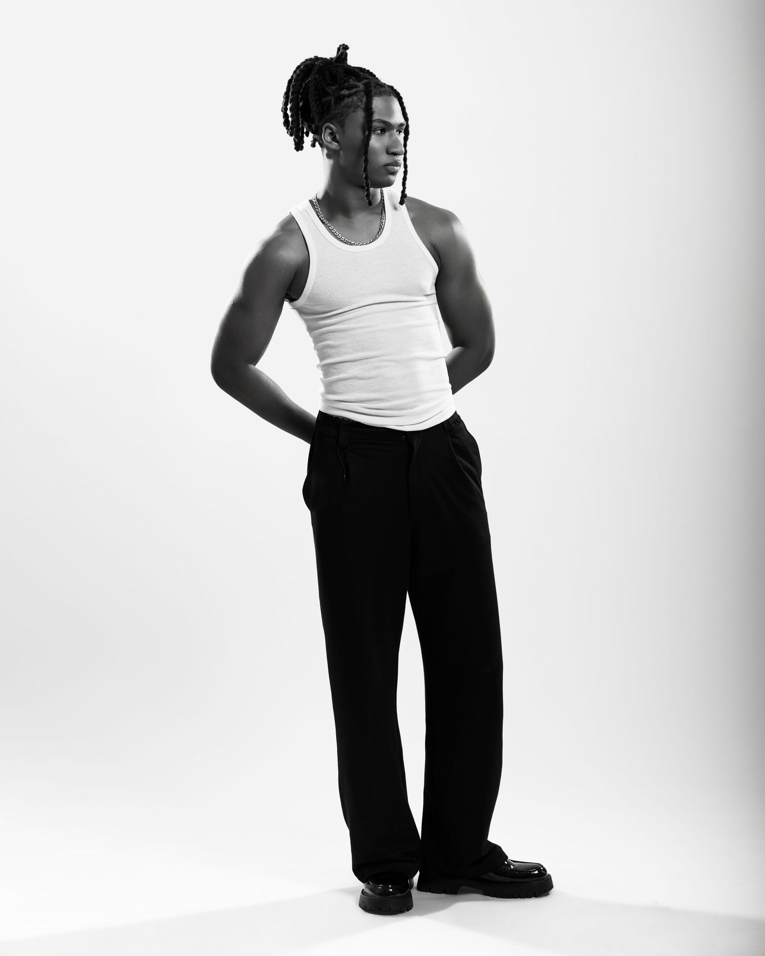 Black and white photo of a young man in a white tank top and black trousers, posing confidently in a studio.