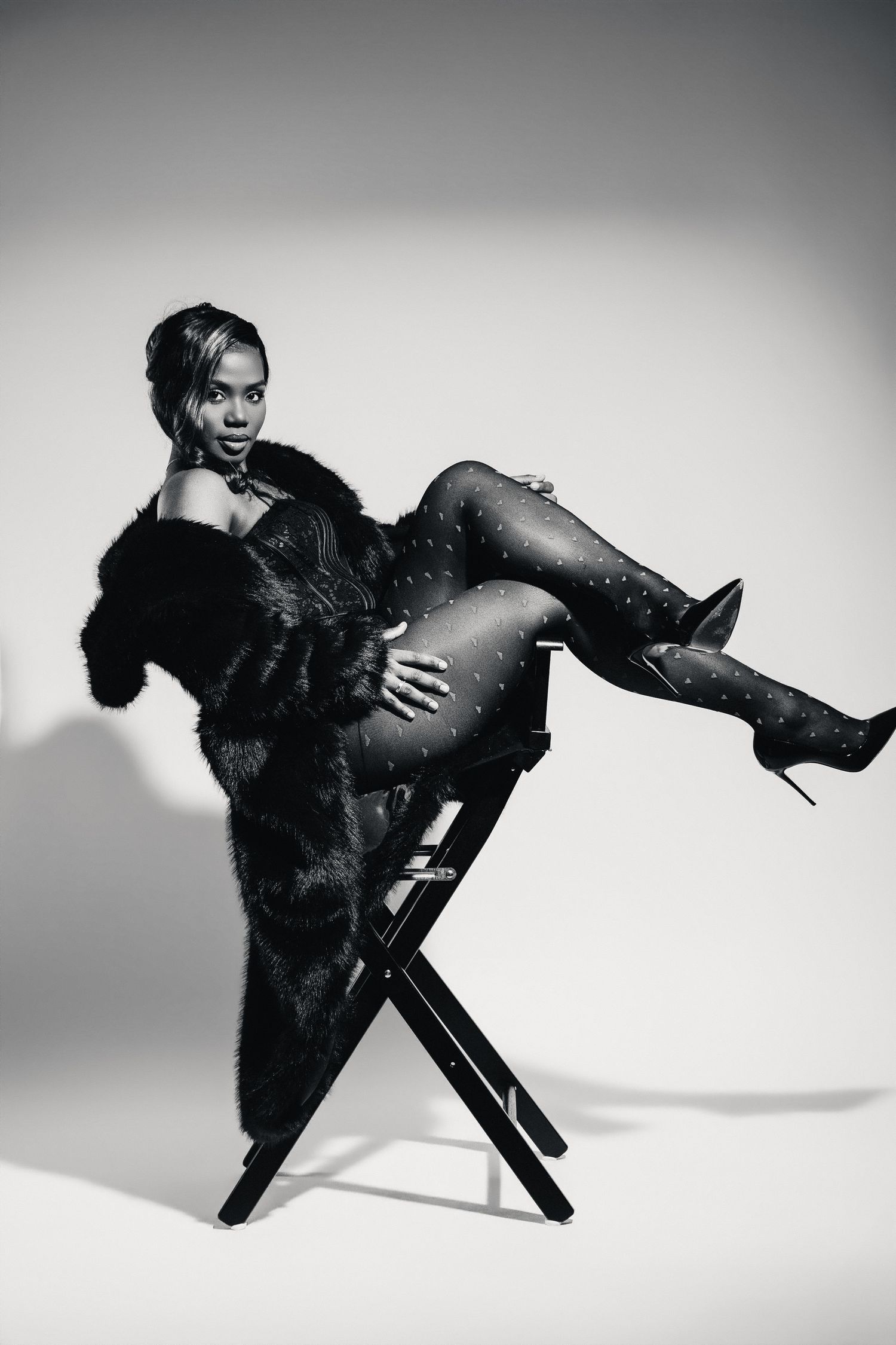 Black and white photo of a woman in fur coat, patterned tights, and heels posing on a director's chair.