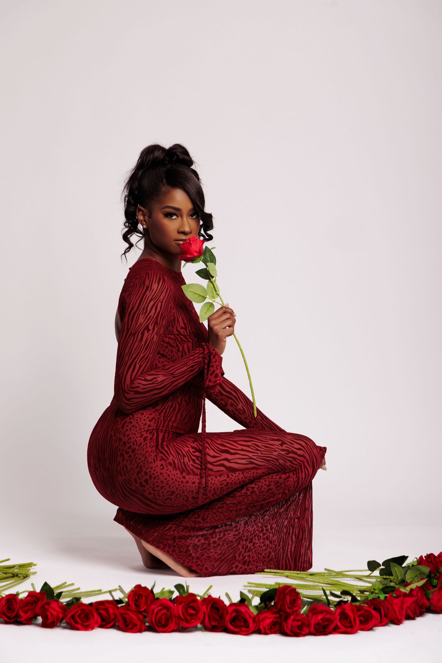 A Black woman in a red lace dress sits gracefully, smelling a red rose, surrounded by roses on a white background.