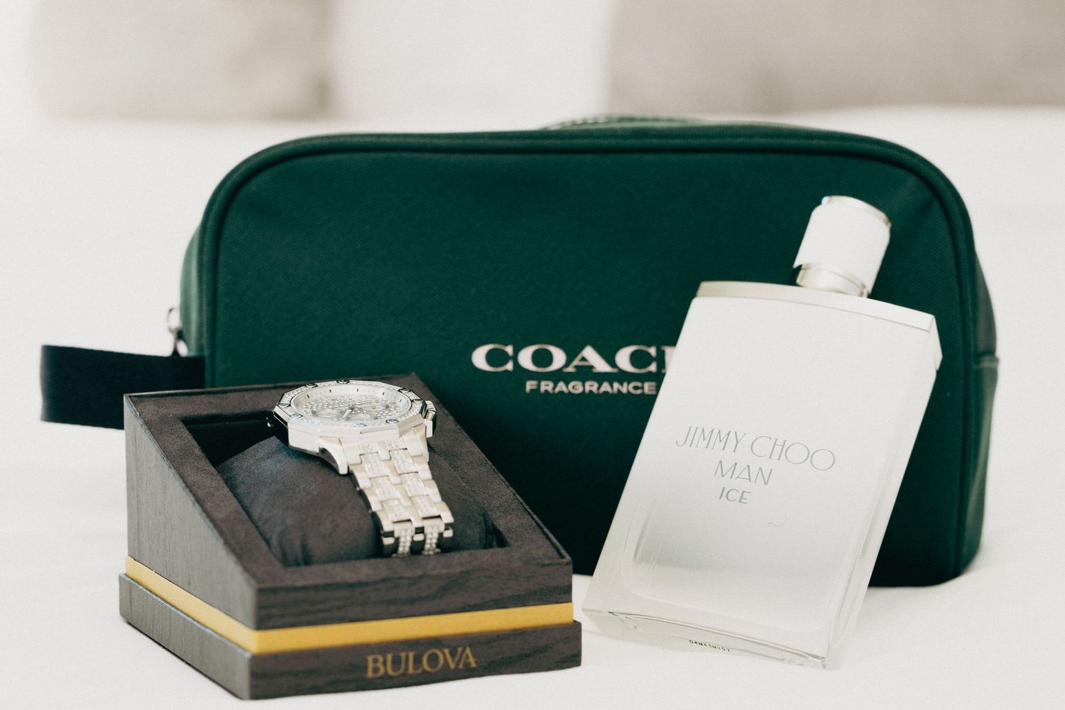 Bulova watch in box beside Jimmy Choo Man Ice cologne and green Coach Fragrance toiletry bag on white surface.