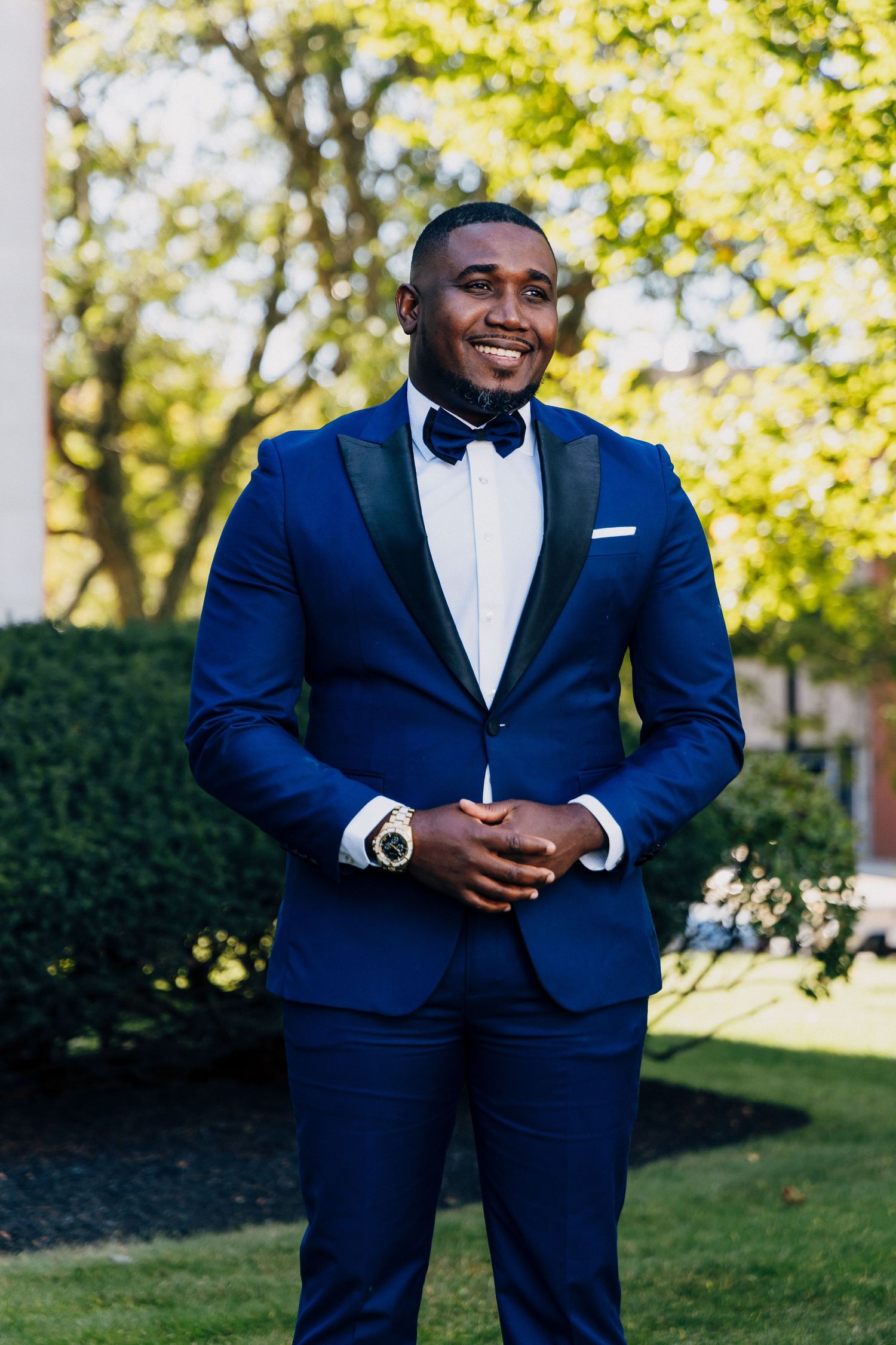 A smiling groom in a navy blue tuxedo with black bow tie stands outdoors on a sunny day.
