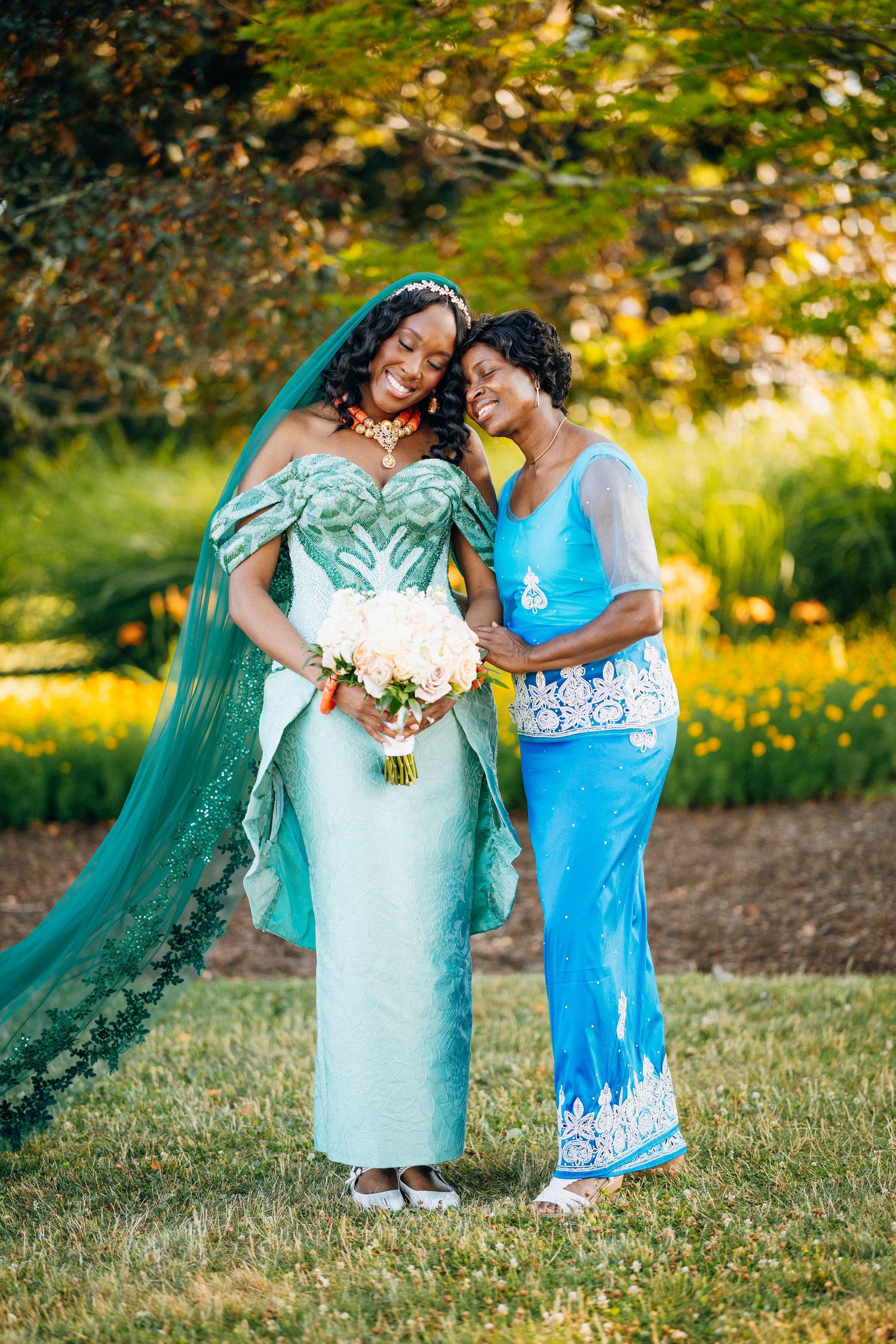 Bride in teal Nigerian wedding dress with long veil poses with older woman in blue lace gown outdoors.