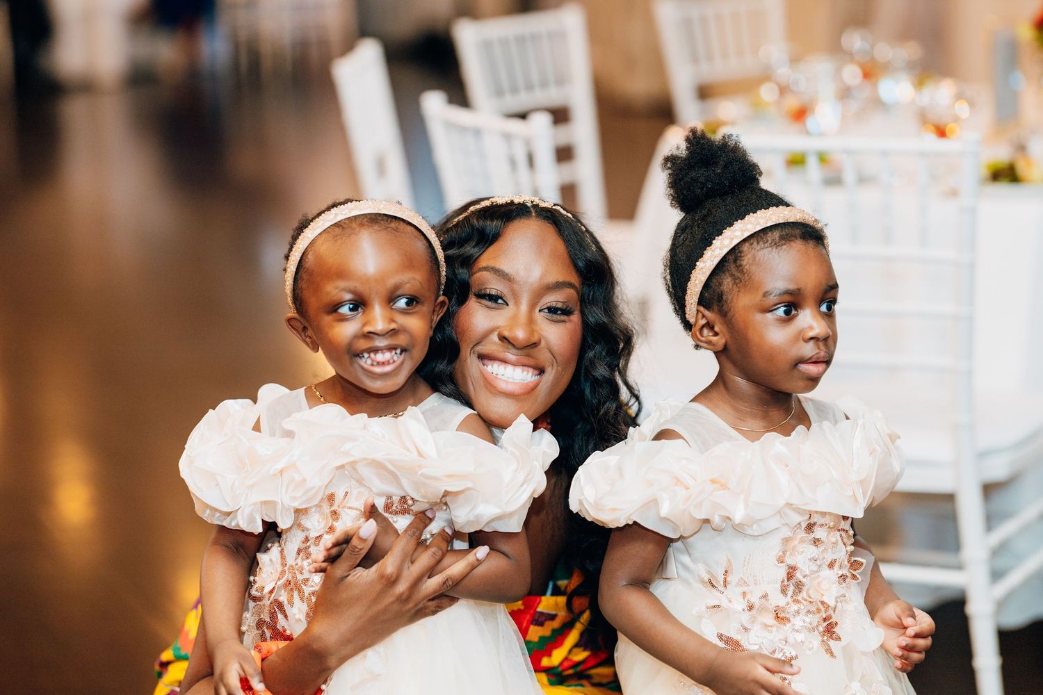 Smiling woman in kente cloth hugs two young girls in white floral dresses at a wedding reception.