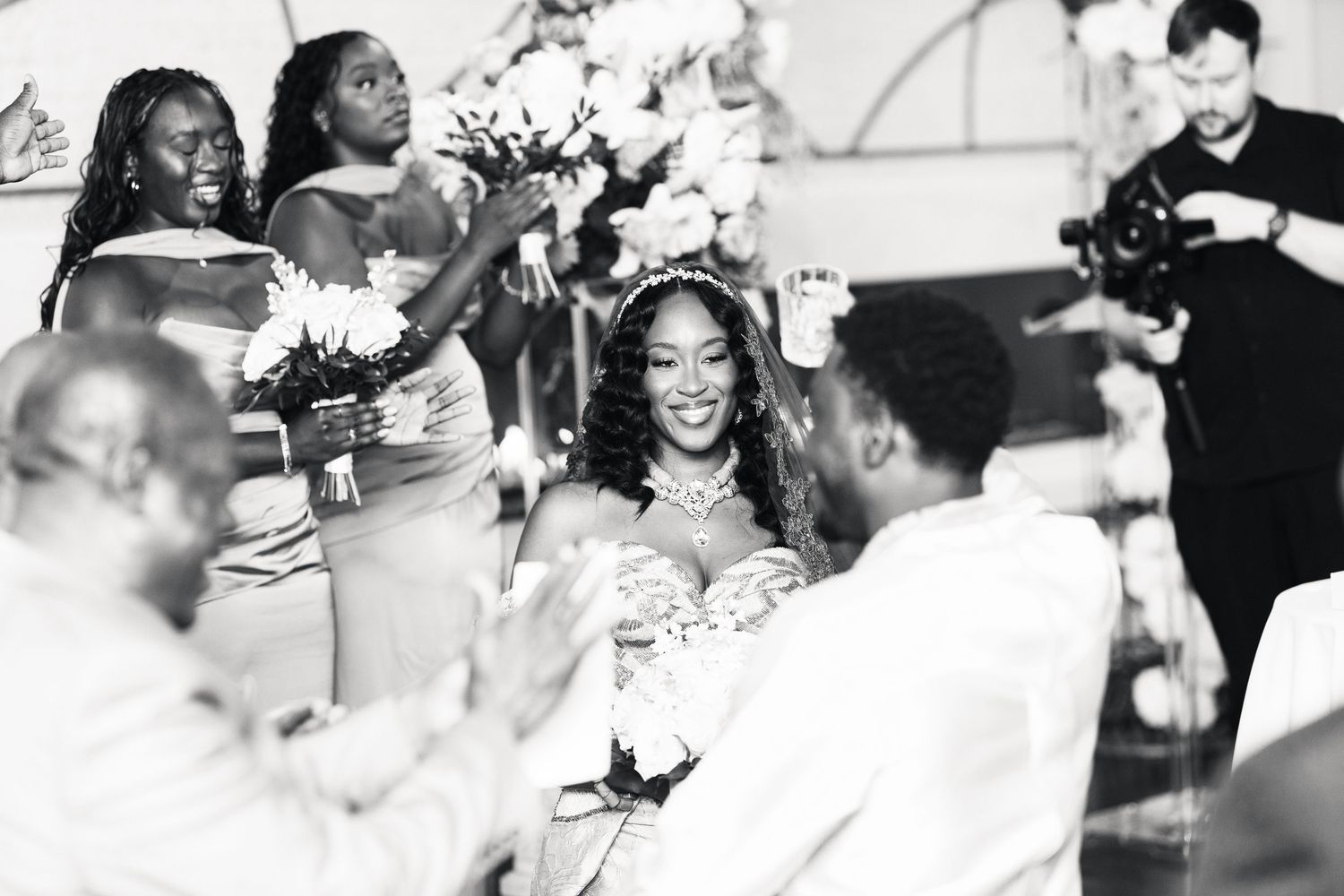 Black and white photo of smiling bride surrounded by bridesmaids holding bouquets at wedding reception.